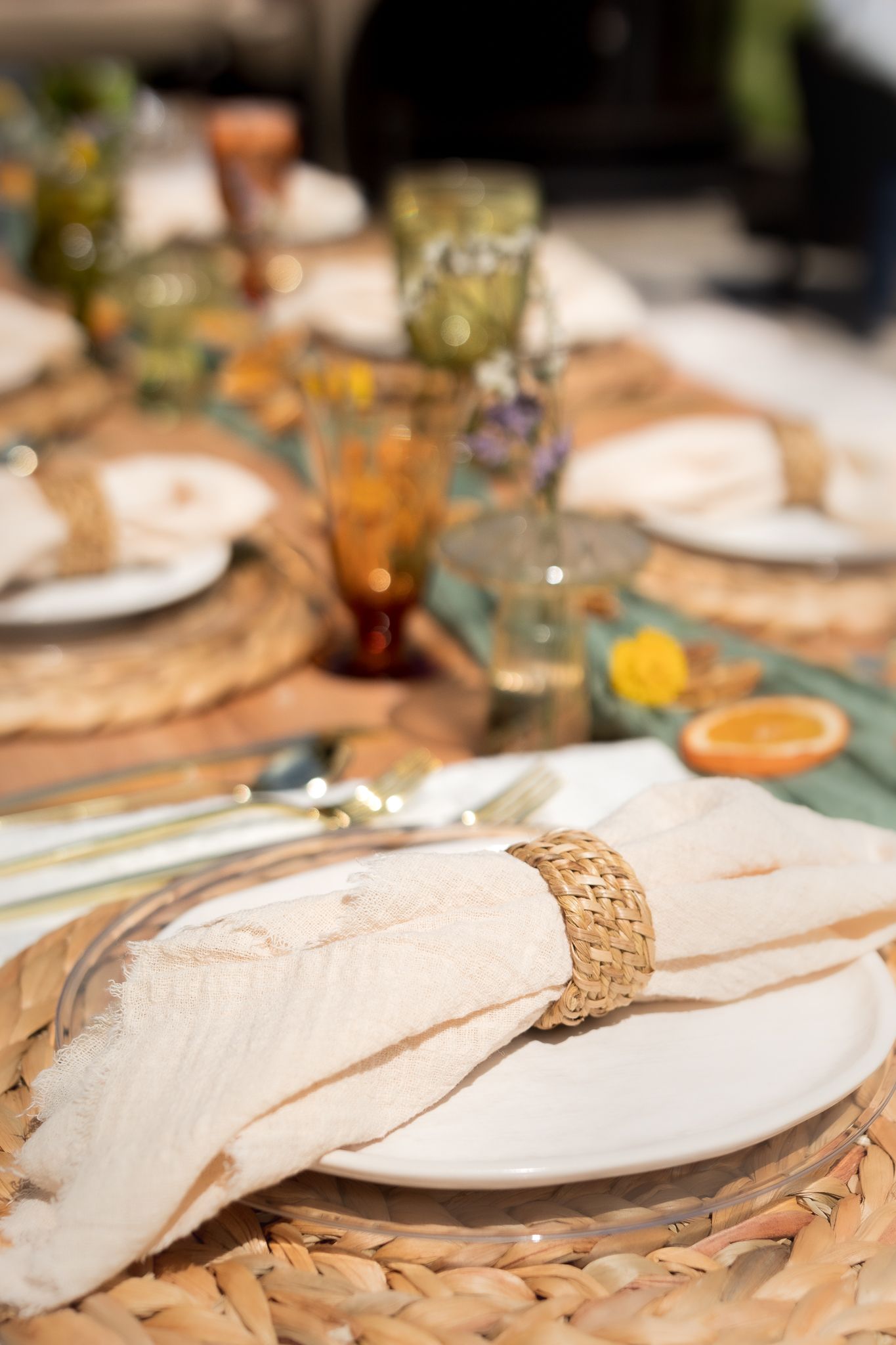 Elegant outdoor table setting with white plates, a woven napkin ring, linen napkin, colorful glasses, and wildflowers on woven chargers.