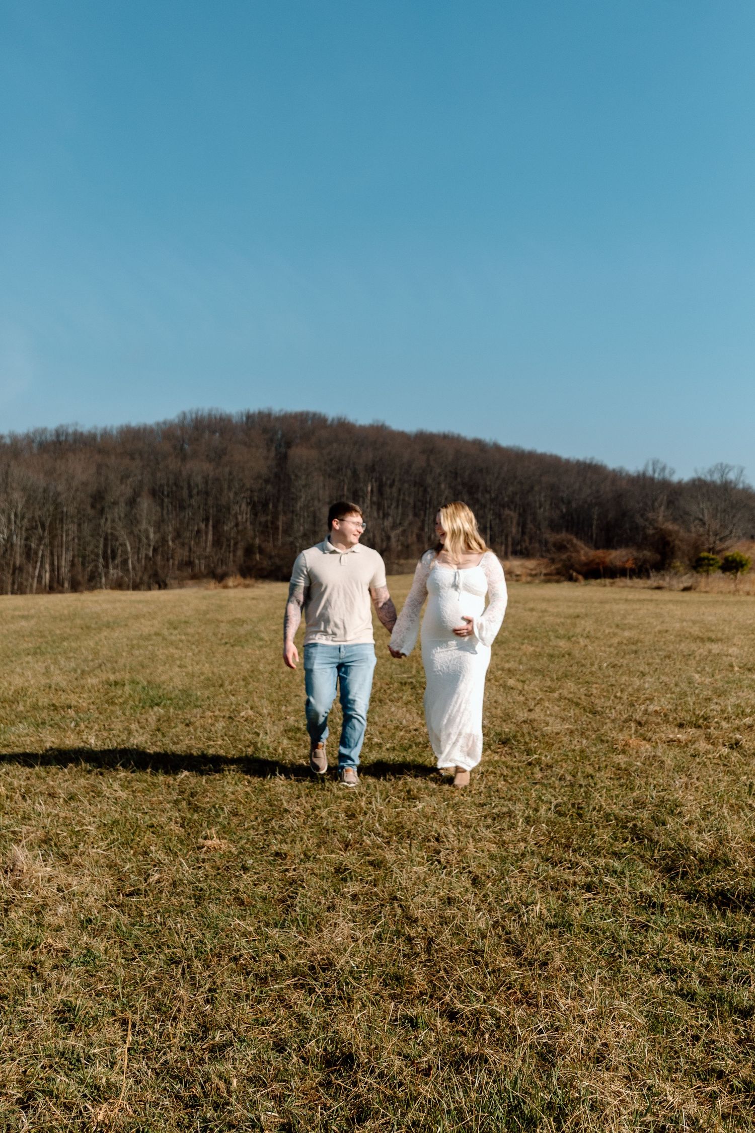 Couple holding hands and walking through a sunny grassy field with a wooded hillside and clear blue sky behind them.