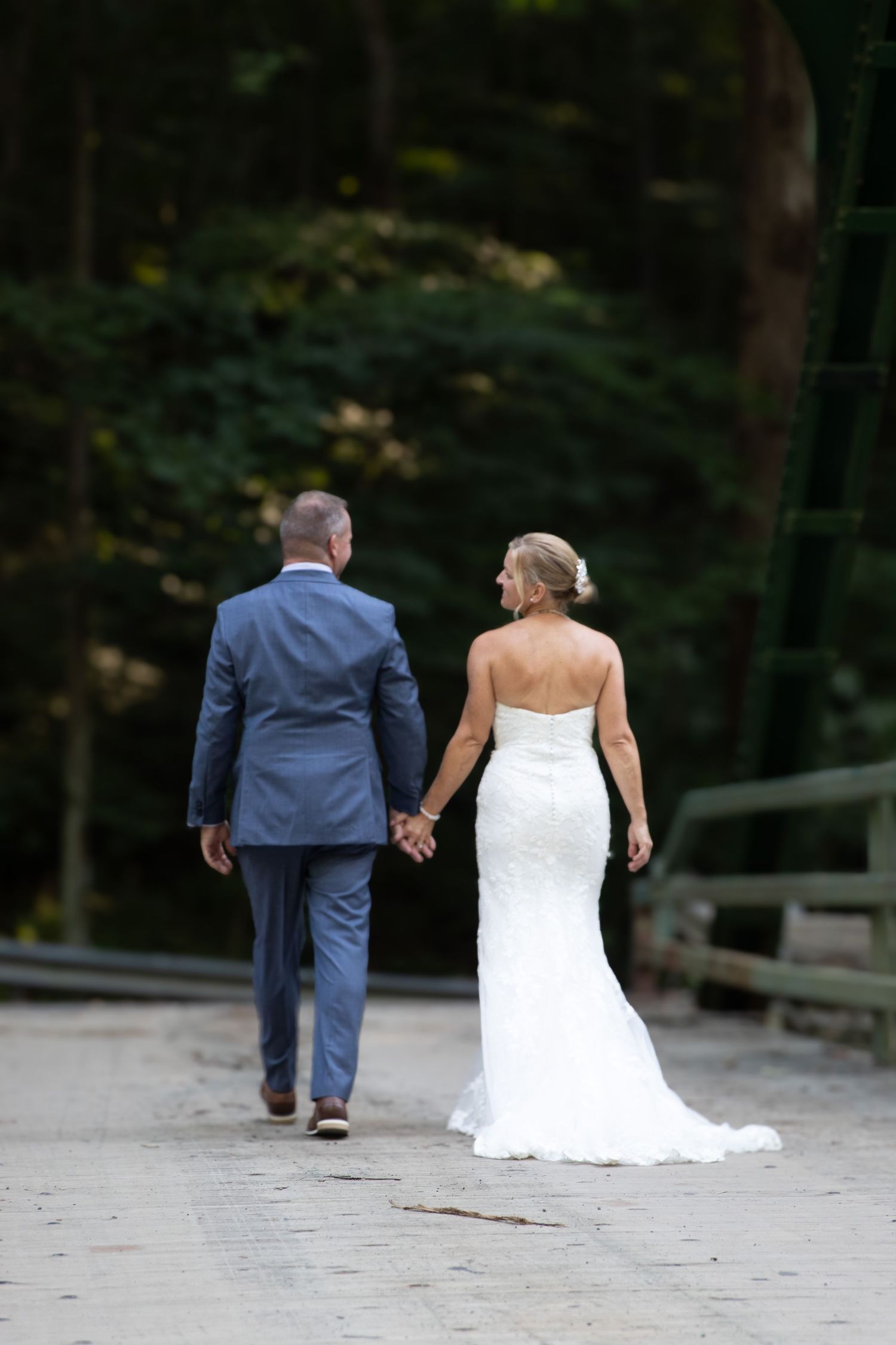 Bride in a white lace gown and groom in a blue suit walk hand in hand along a wooded path after their wedding ceremony.