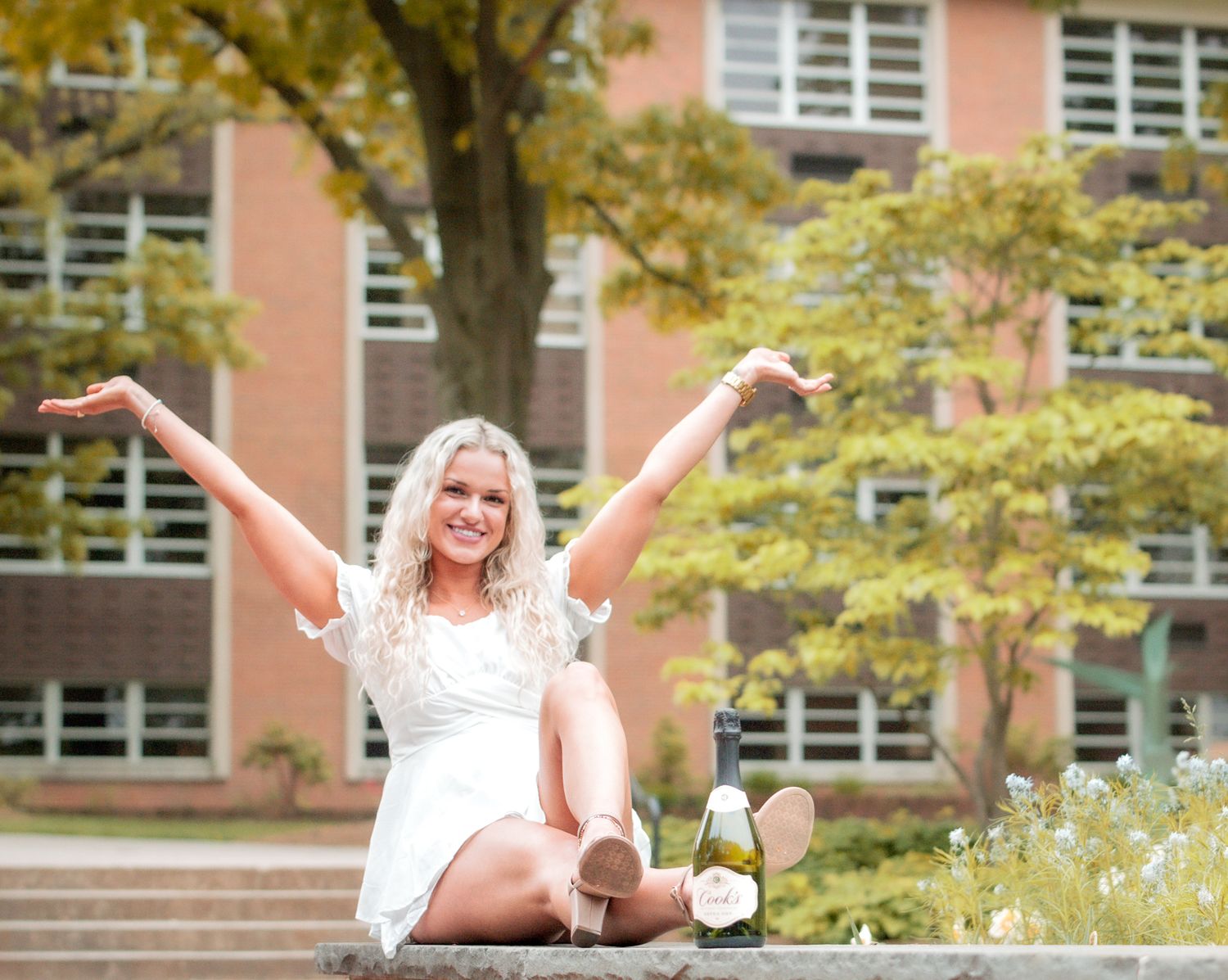 Smiling woman in a white dress sits on campus steps with arms raised, a champagne bottle beside her, brick building and autumn trees behind.