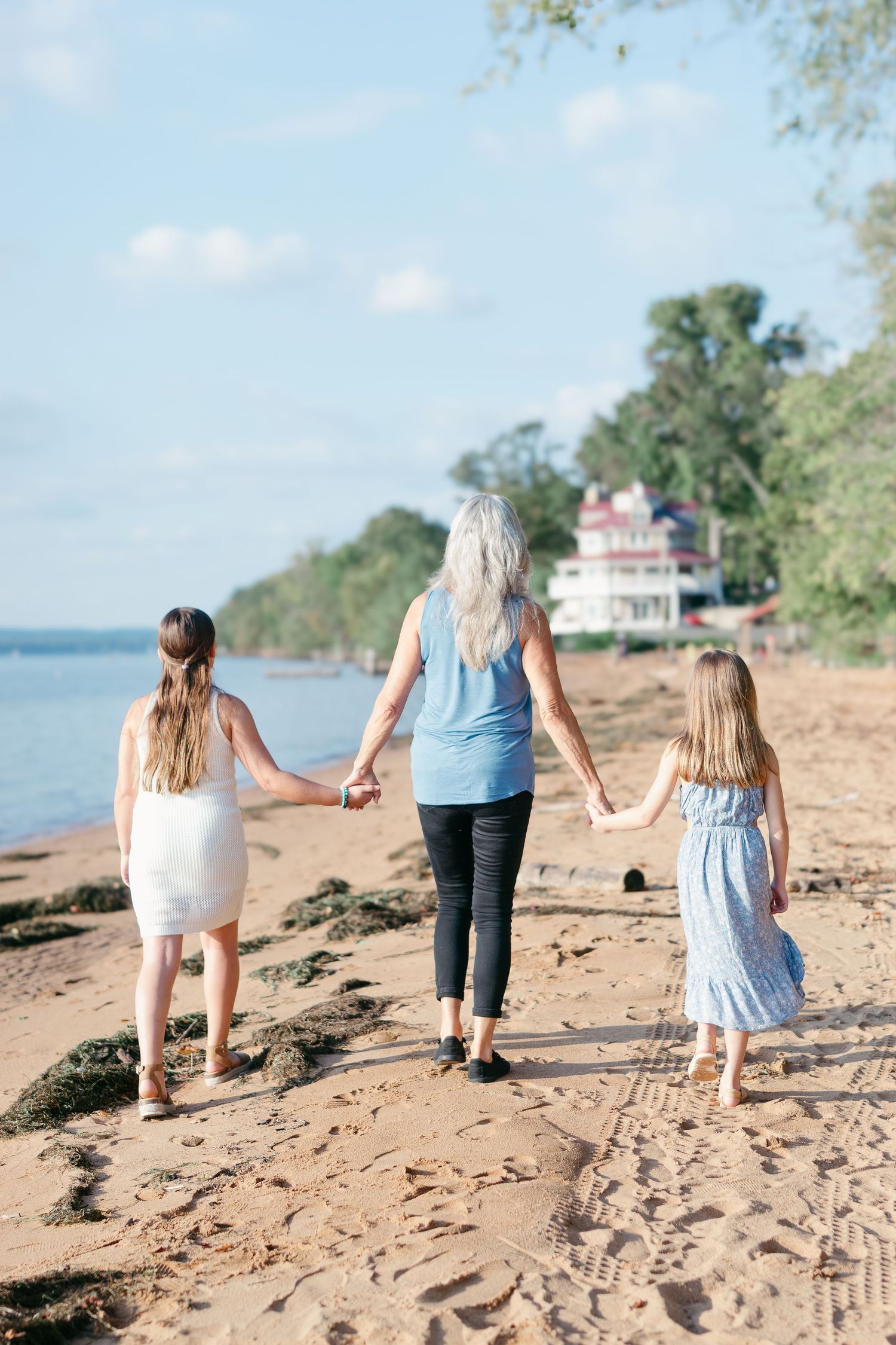A woman and two young girls hold hands and walk along a sandy beach toward a distant house, with calm water and trees nearby.