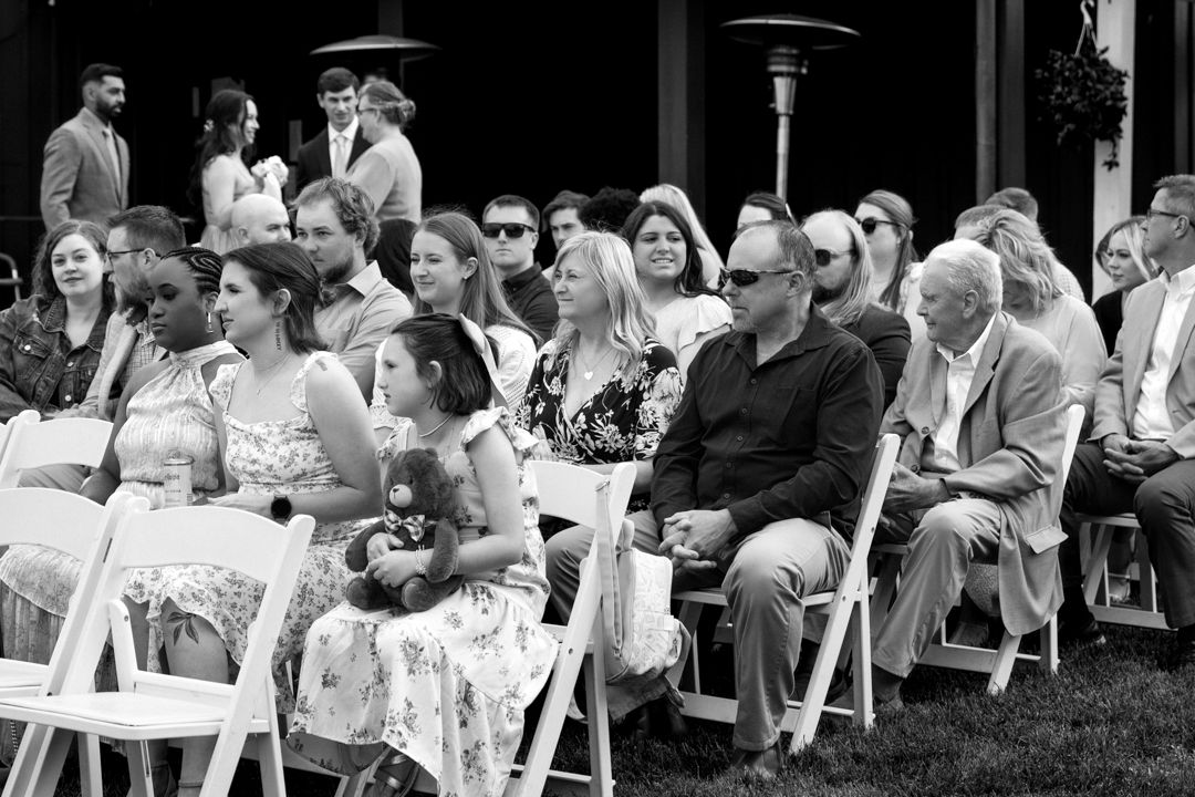 Wedding guests seated in white folding chairs on a lawn watch an outdoor ceremony, including a young girl holding a stuffed animal.