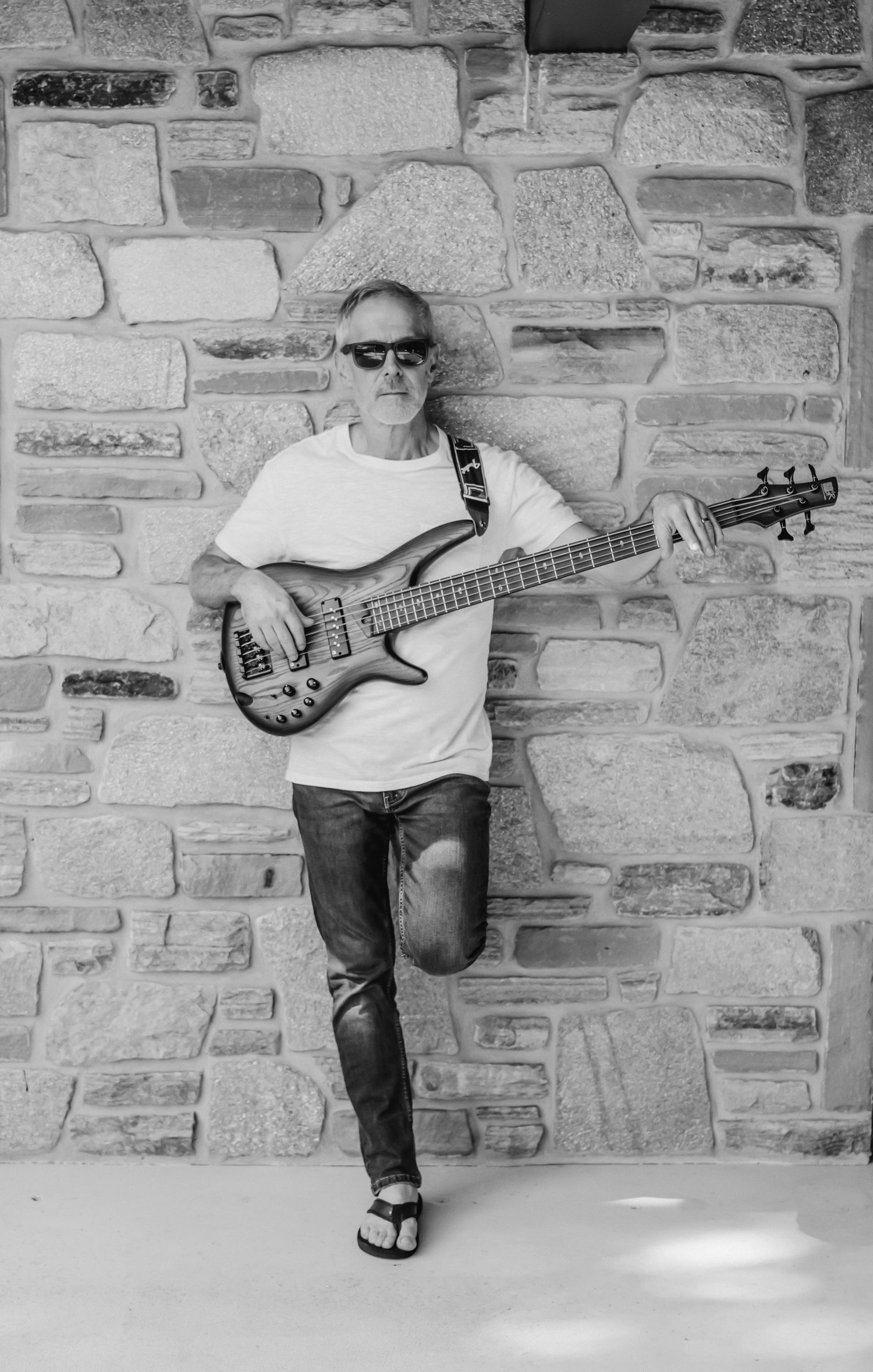Black and white photo of a man in sunglasses and a white t-shirt playing a bass guitar while leaning against a stone wall.