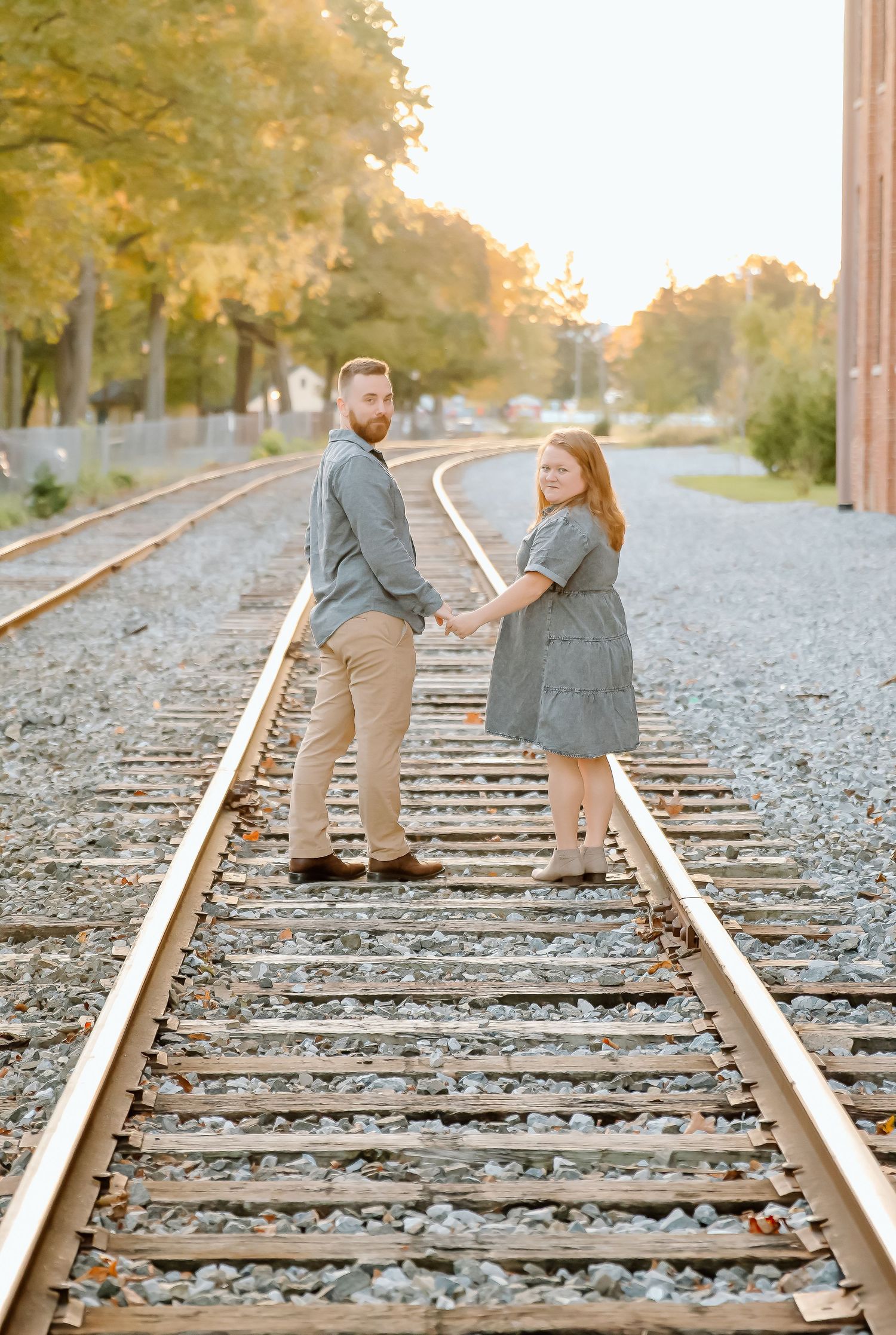 Couple holding hands and looking back over their shoulders while standing on railroad tracks at sunset during an engagement photo session.