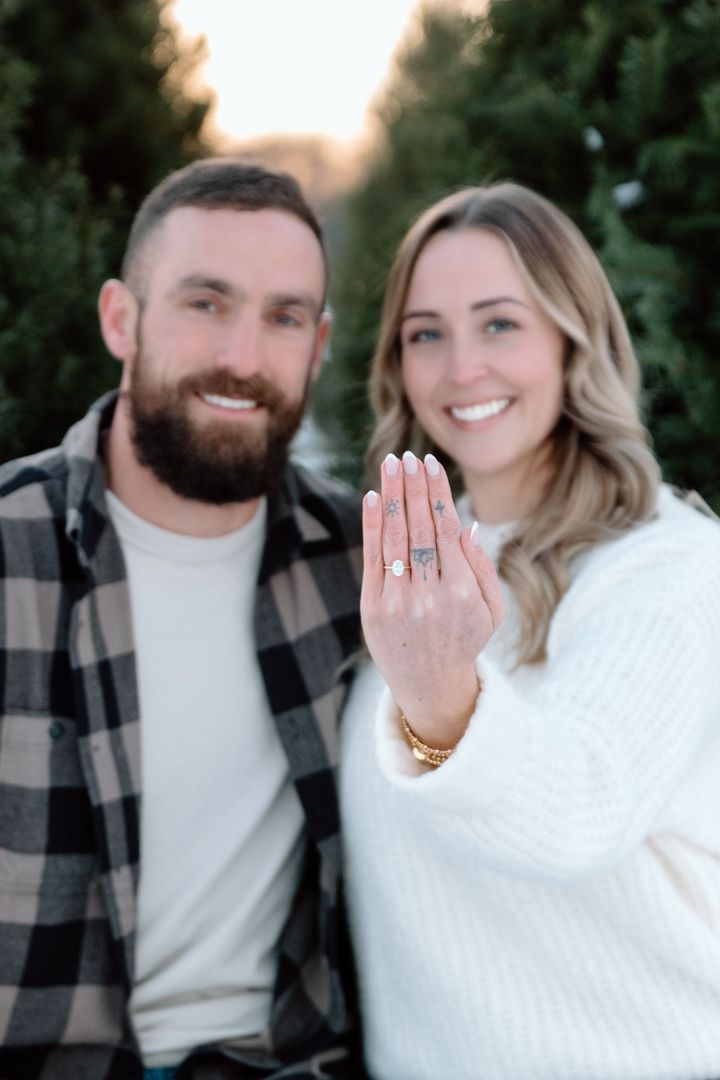 Smiling couple outdoors near evergreen trees, with the woman holding up her hand to show off a sparkling diamond engagement ring.