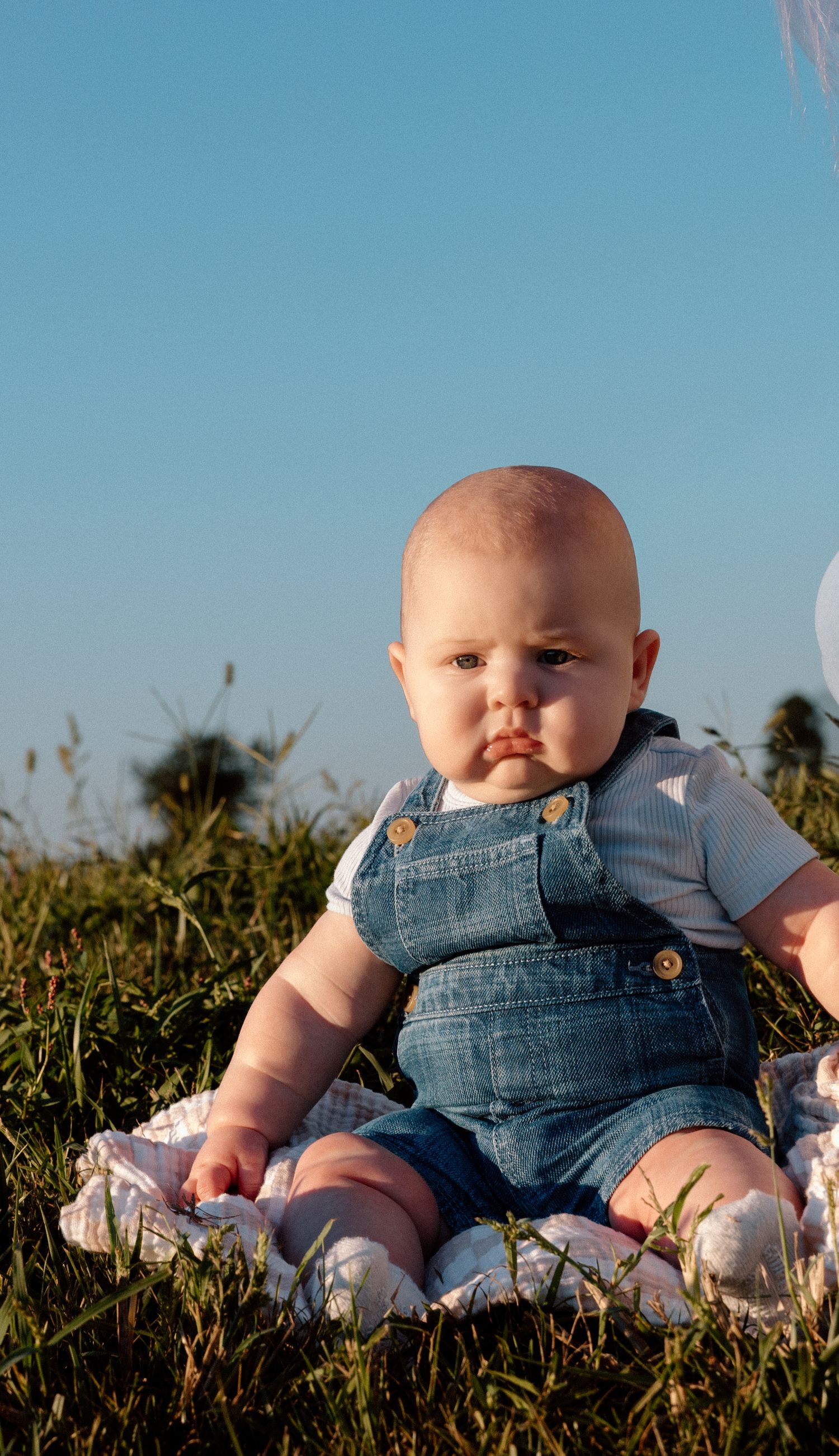 Baby in denim overalls and a white shirt sits on a blanket in a grassy field under a clear blue sky at golden hour.