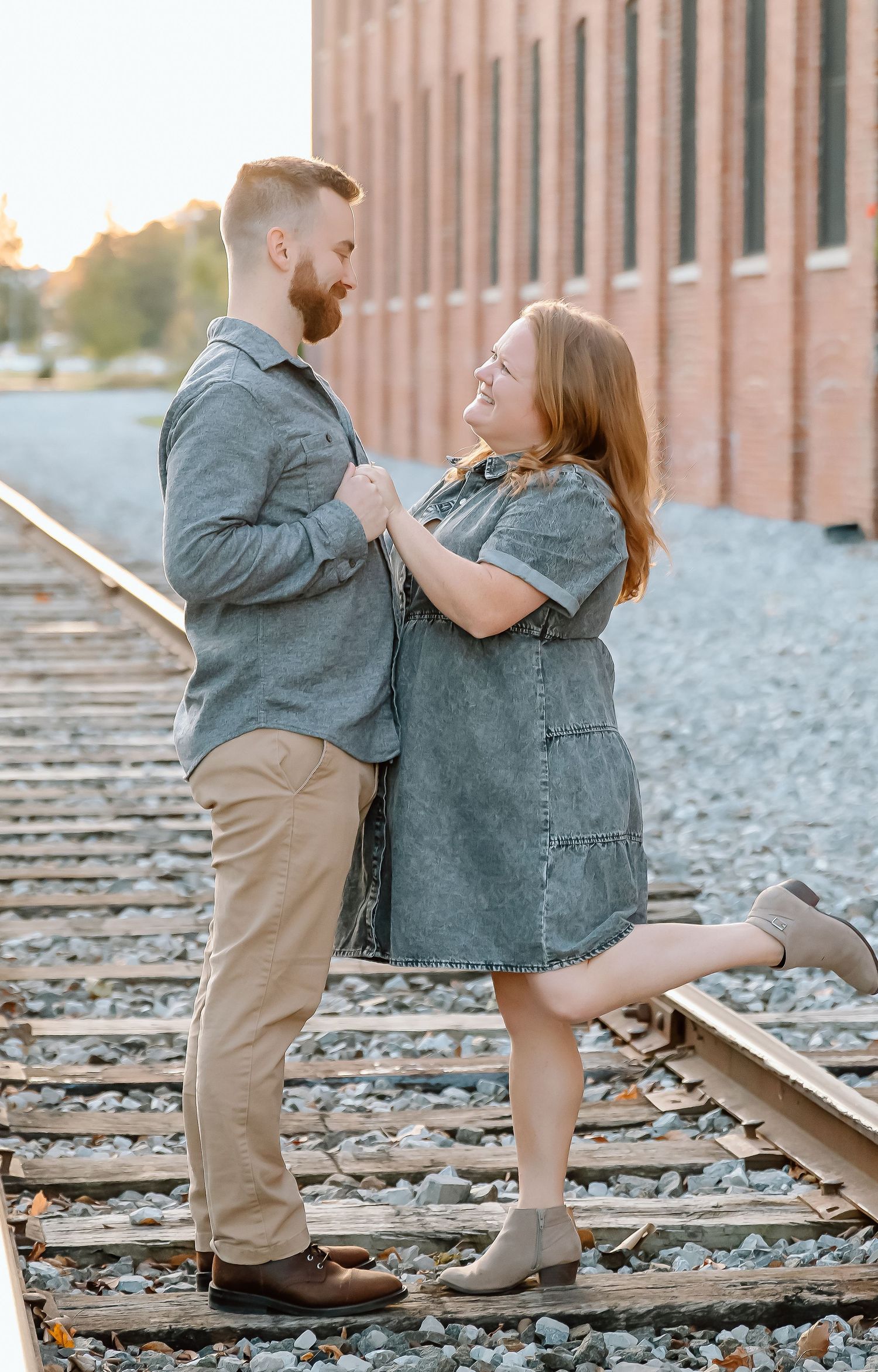 Smiling couple holding hands and embracing on railroad tracks near a brick building during a sunset engagement photo session.