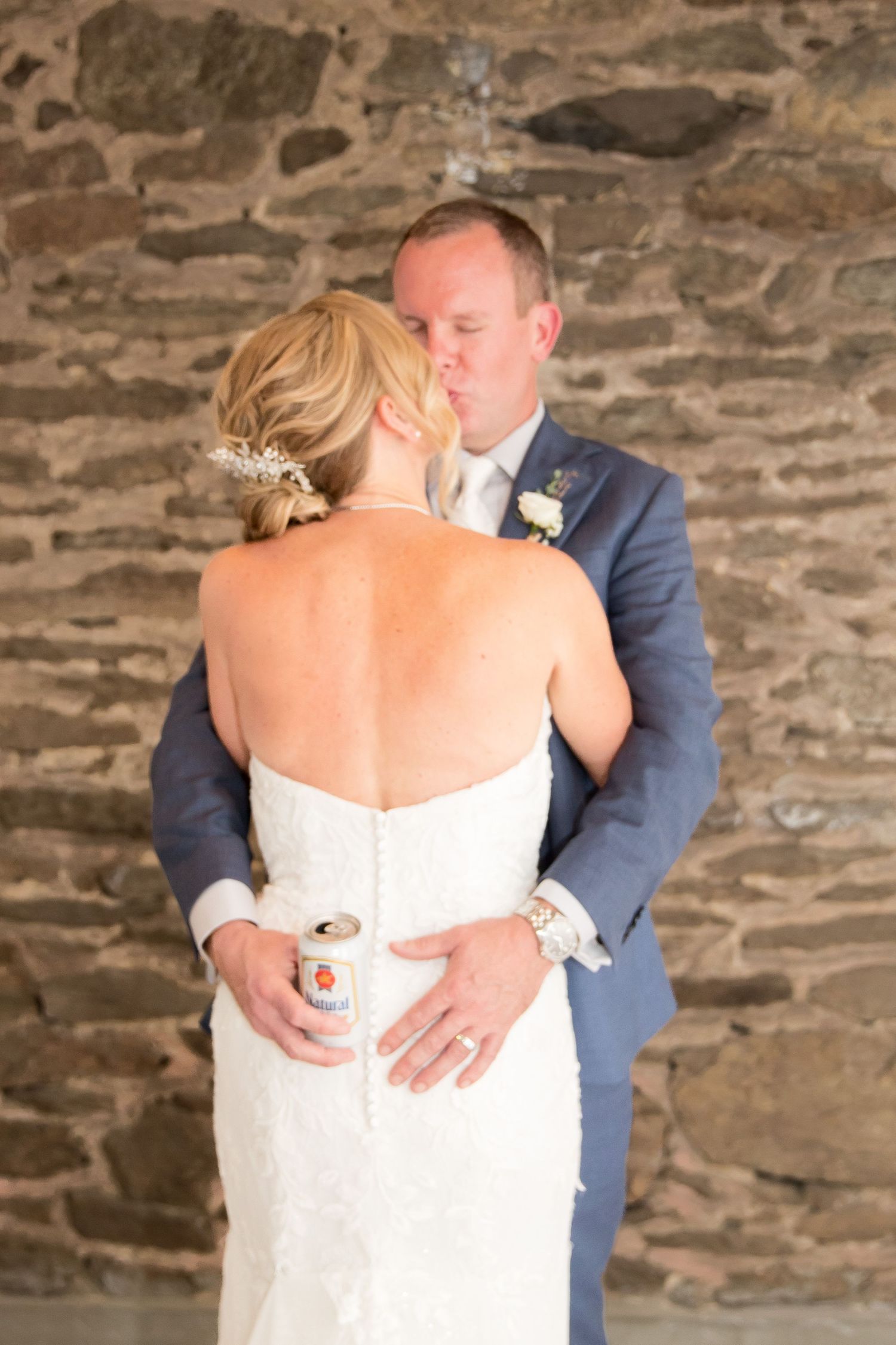 Bride in a strapless white lace gown embraces a groom in a blue suit while holding a beer can against a rustic stone wall.