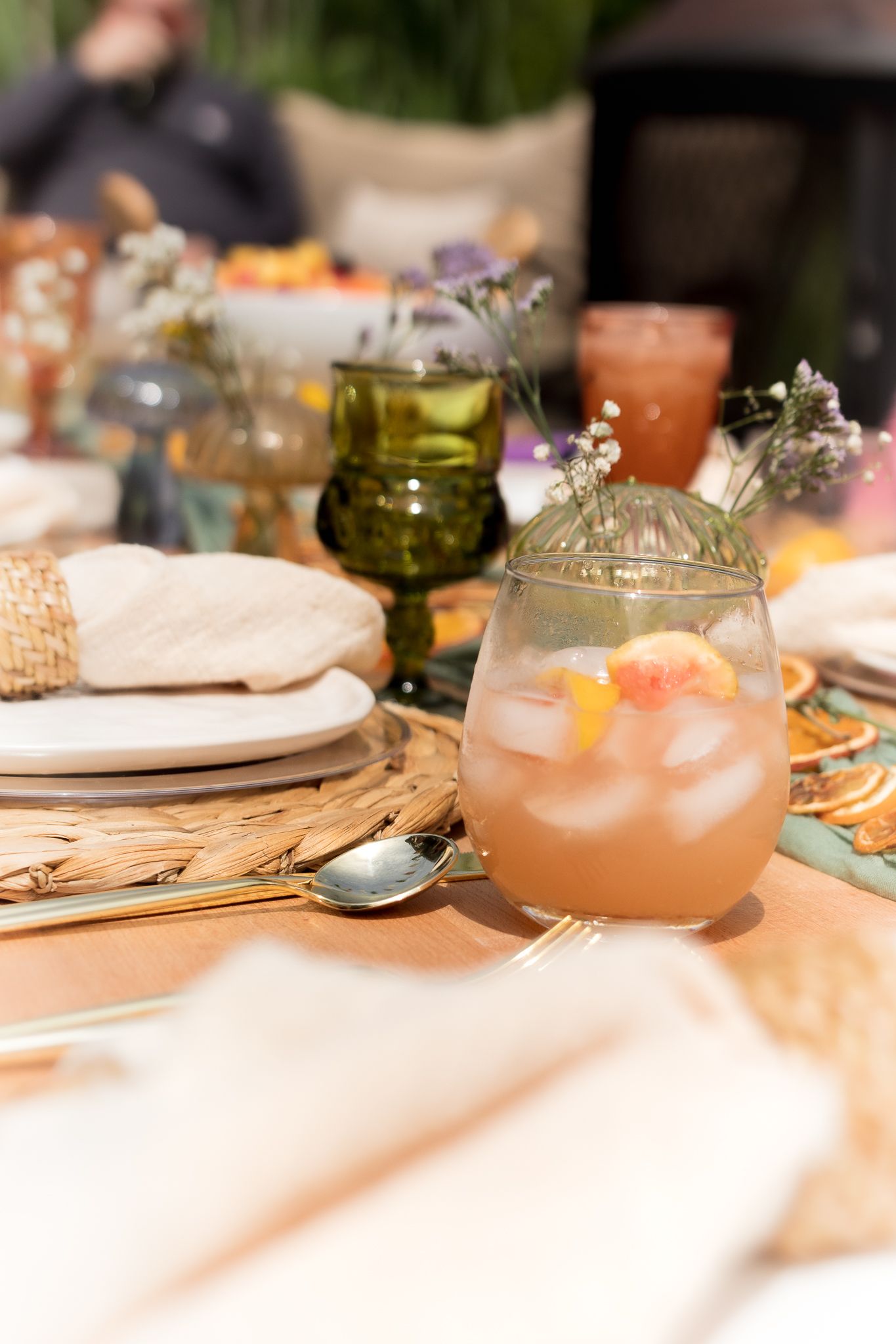 A wedding reception table with a peach cocktail in a glass, flowers, a green goblet, woven placemats and folded linen napkins.