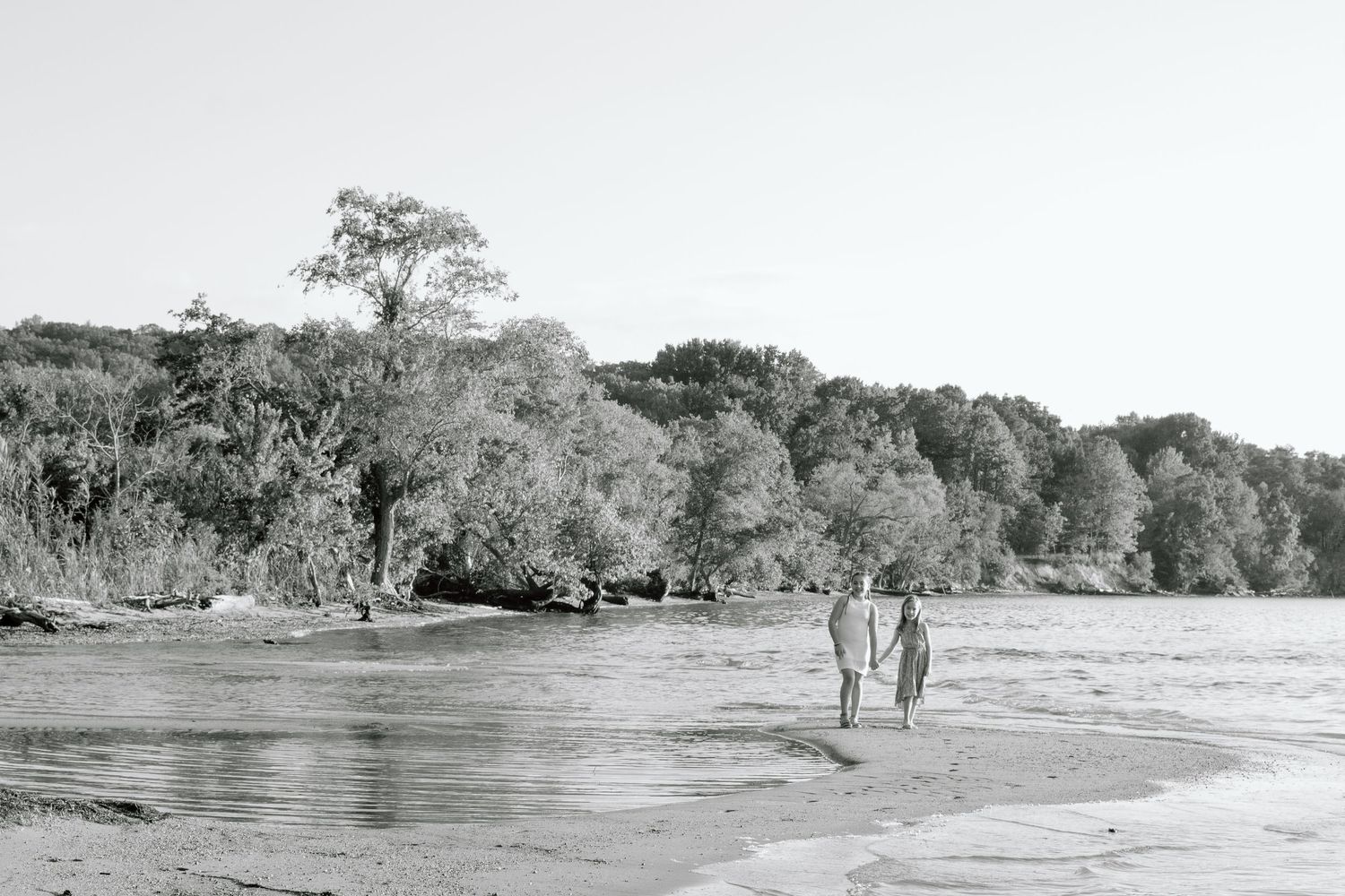 Black and white photo of a couple holding hands while walking along a quiet sandy shoreline with lush trees in the background.