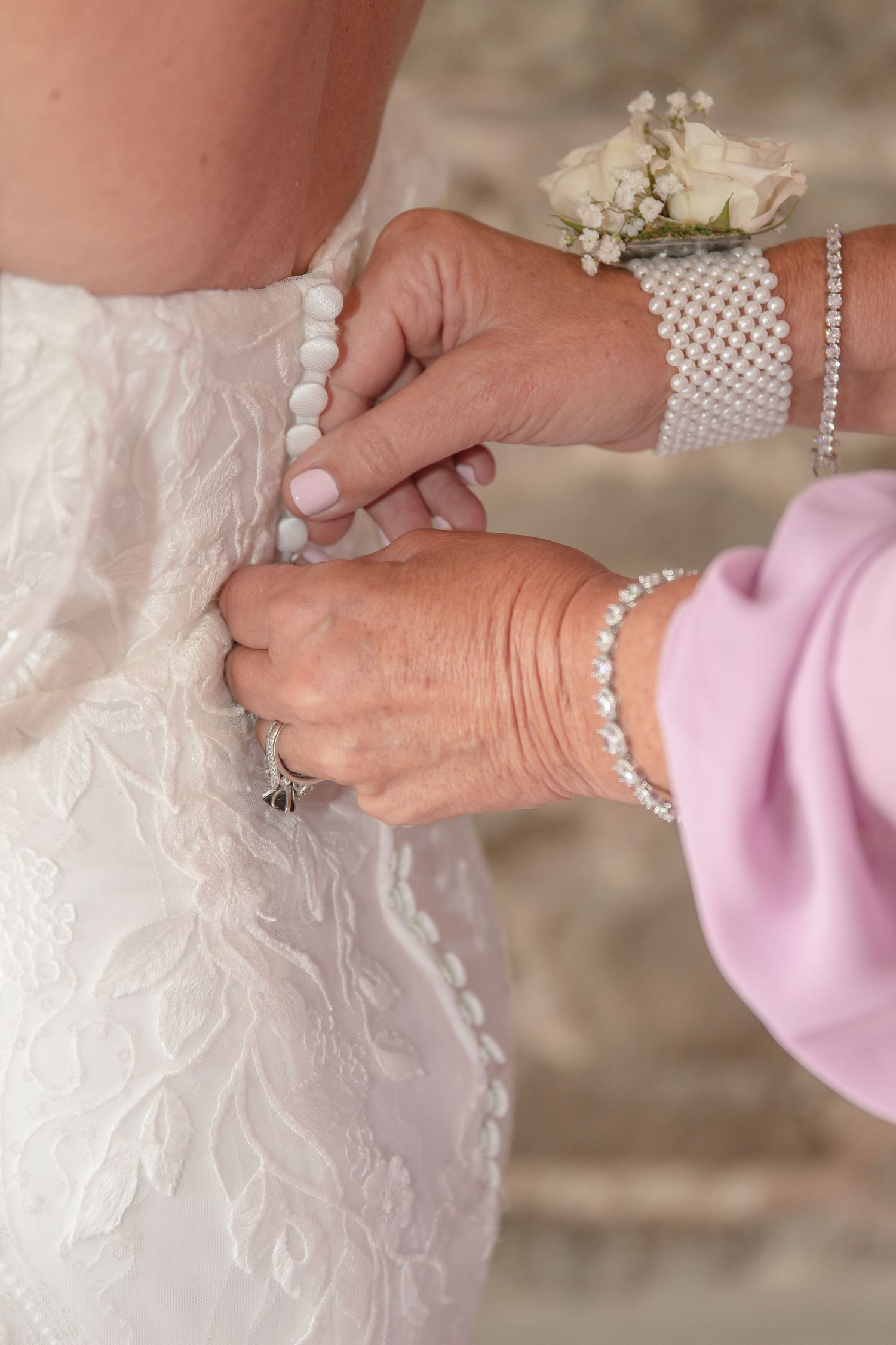 A mother in a pink sleeve with a pearl cuff bracelet buttons the pearl buttons on the back of a lace bridal gown.