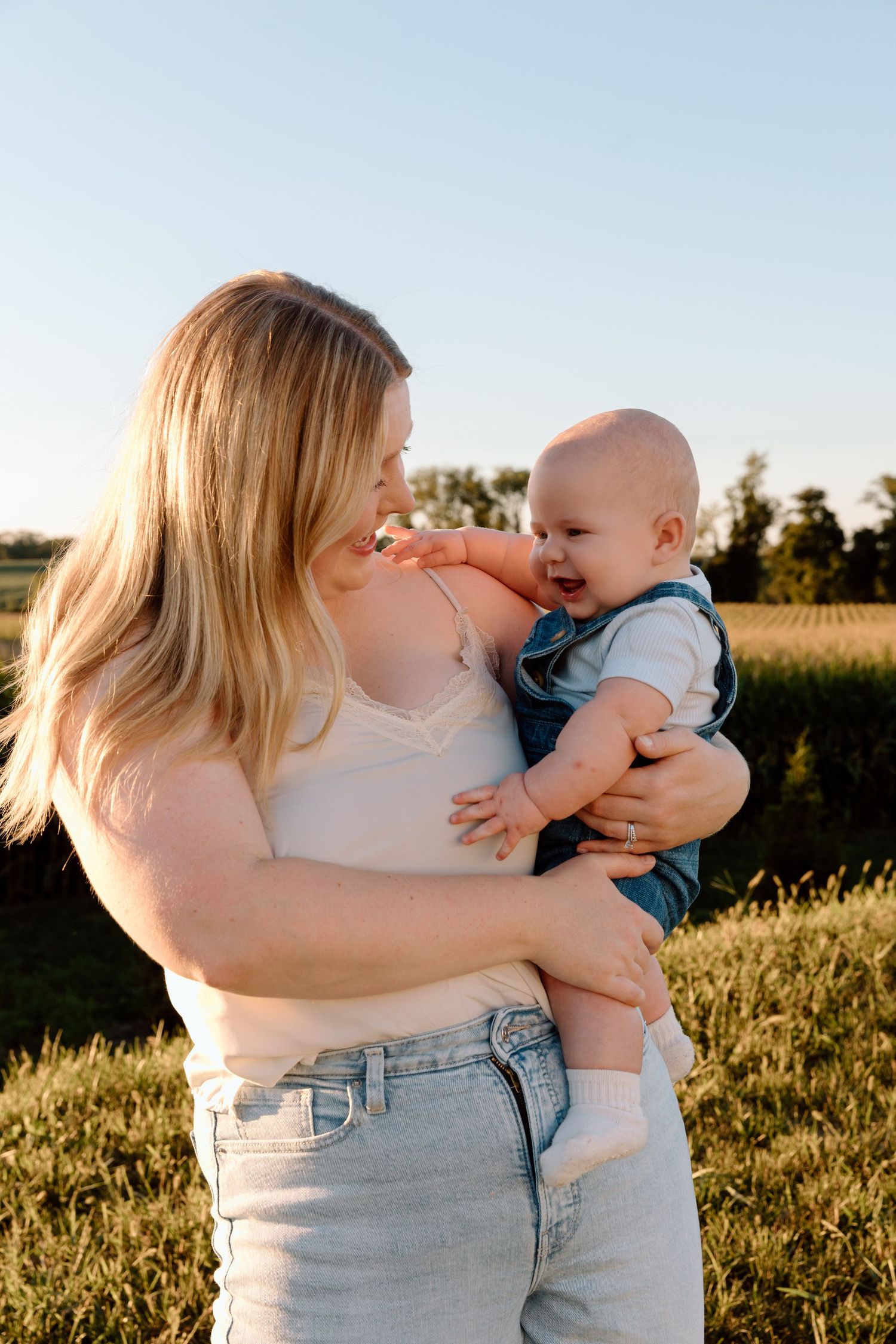 A smiling woman with long blonde hair holds a laughing baby in denim overalls while standing in a sunlit grassy field at golden hour.