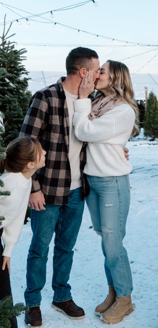 Couple kissing in the snow at a Christmas tree farm while a young girl in a white sweater looks on, with string lights overhead.
