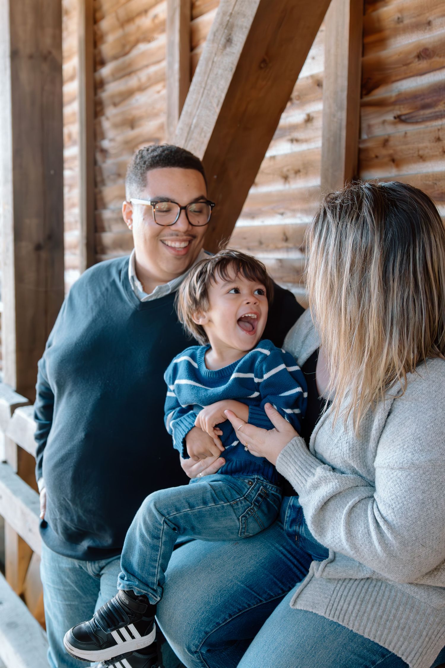 A smiling man with glasses and a laughing woman hold a giggling young boy in a striped sweater against a rustic wooden barn wall.