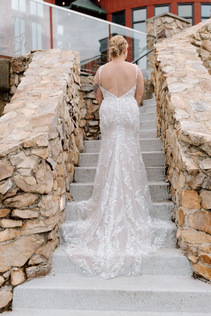 A bride in a lace gown with a long train stands on a rustic stone staircase, viewed from behind.