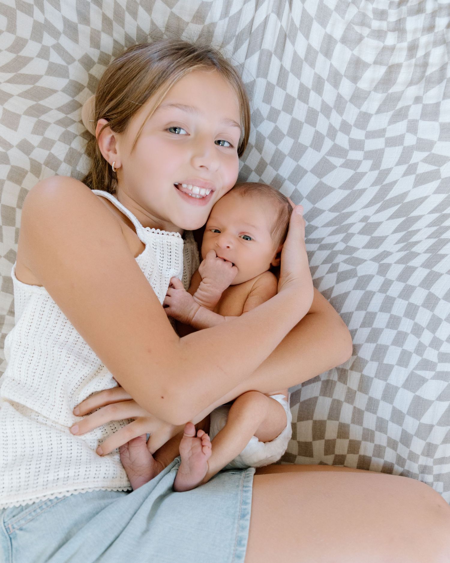 A smiling girl in a white knit top lies on a checkered blanket, gently cuddling a sleeping newborn baby against her cheek.
