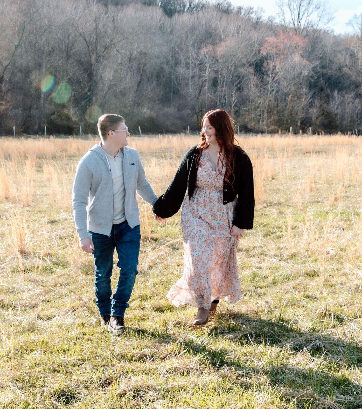 Couple holding hands and walking through a golden grassy field with autumn trees in the background during an engagement session.