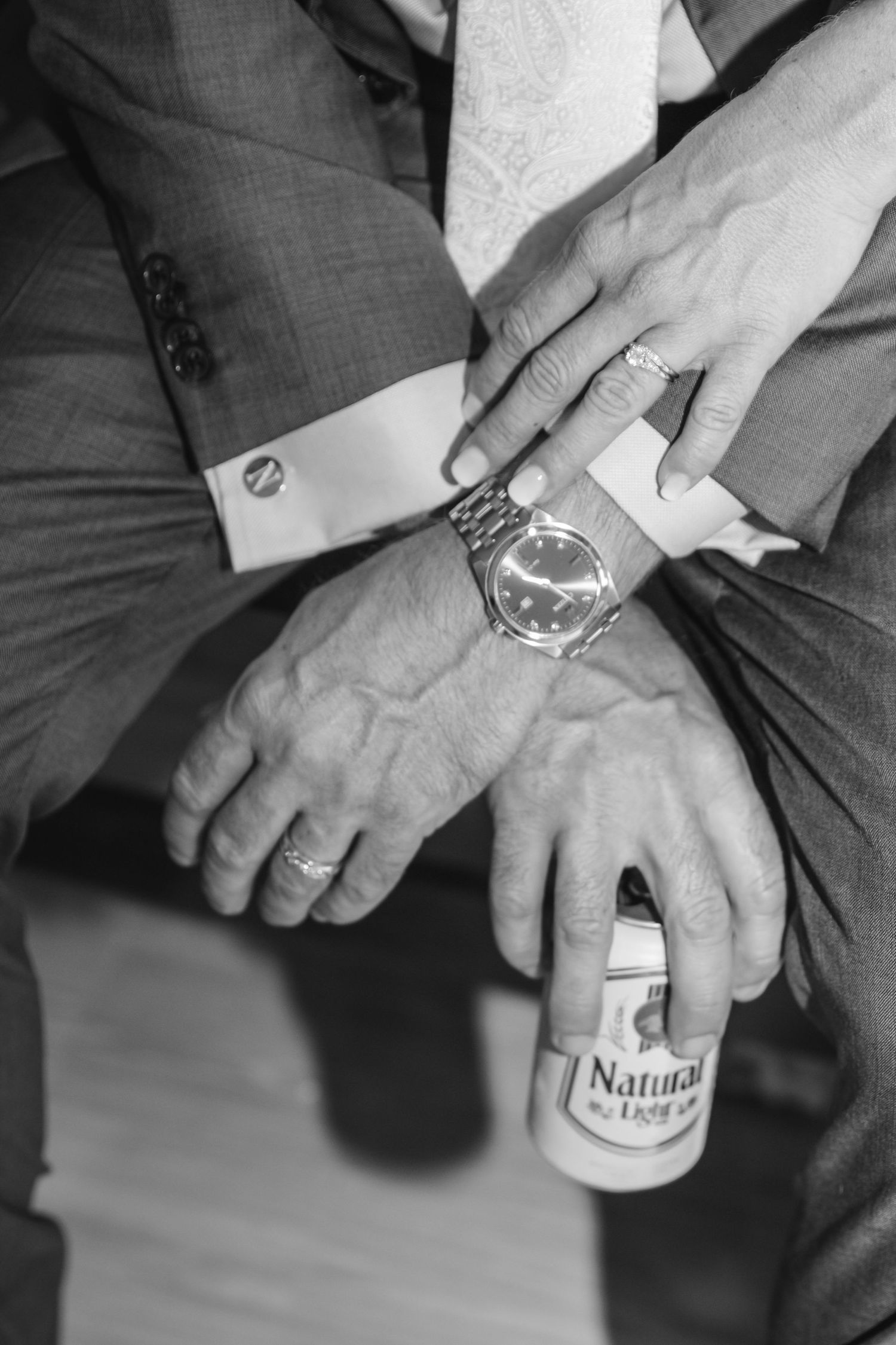 Black and white close-up of a couple's hands with wedding rings, a wristwatch, cufflink and a can of beer.