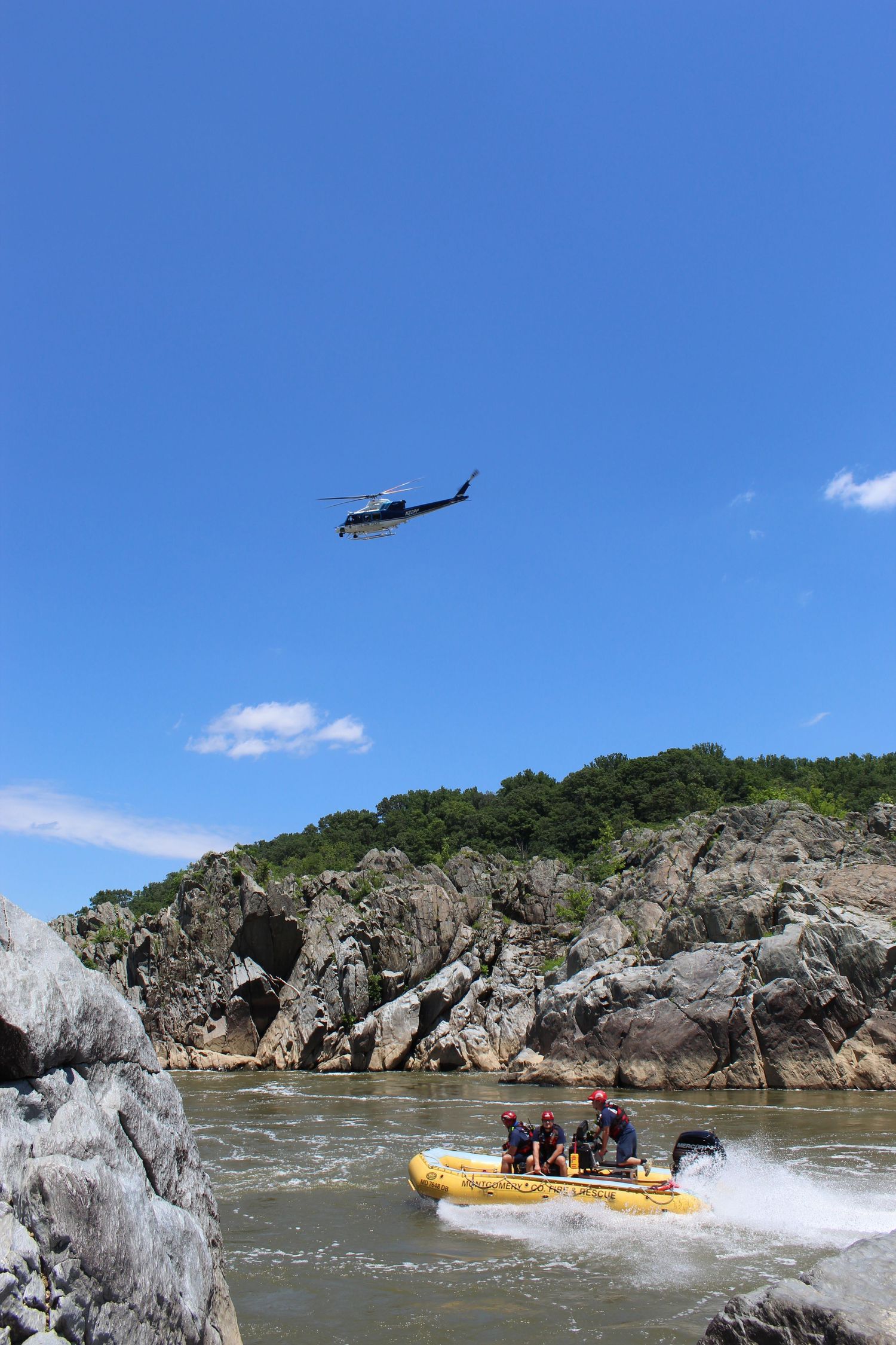 Helicopter flies over a yellow inflatable rescue raft with crew on rushing river rapids beside rocky cliffs under a clear blue sky.