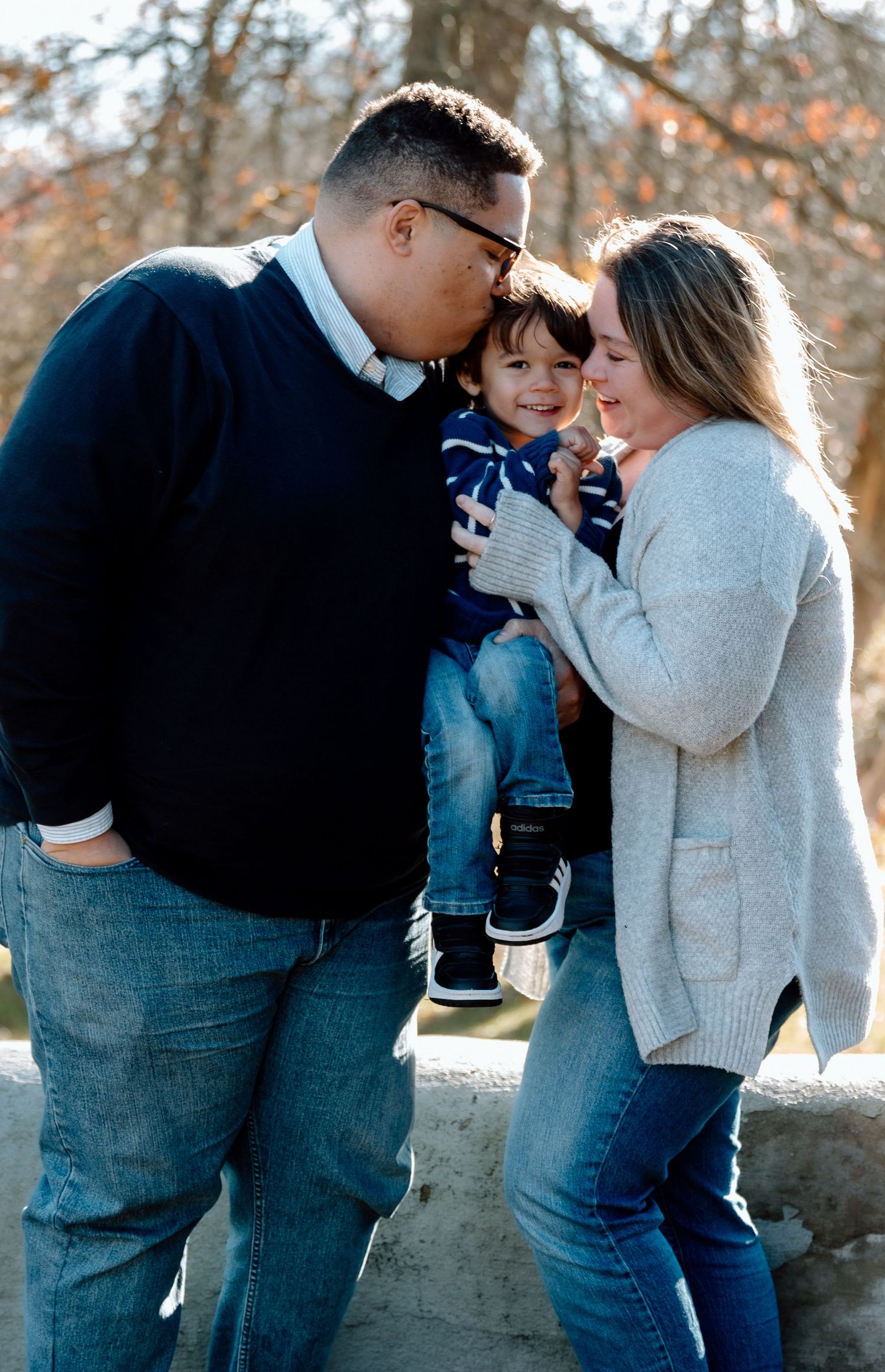 A smiling family of three poses outdoors as a father kisses his young son's cheek while the mother holds the child near a stone wall.