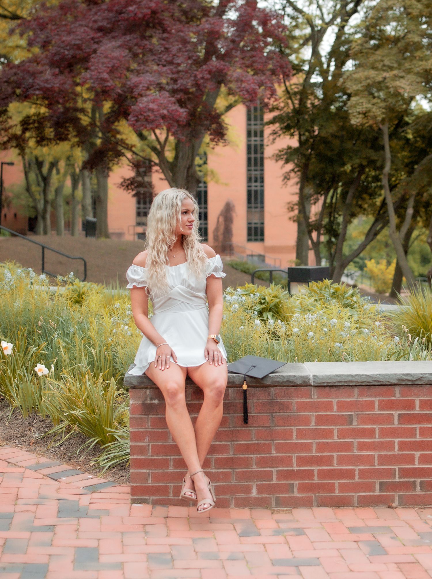 A young woman with long wavy blonde hair in a white dress sits on a brick wall with a graduation cap in a garden on campus.