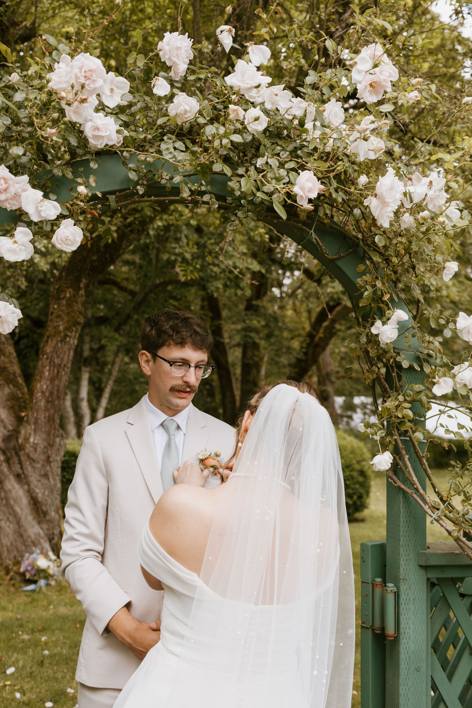 Groom in beige suit stands with bride under floral arch of white roses at outdoor ceremony.