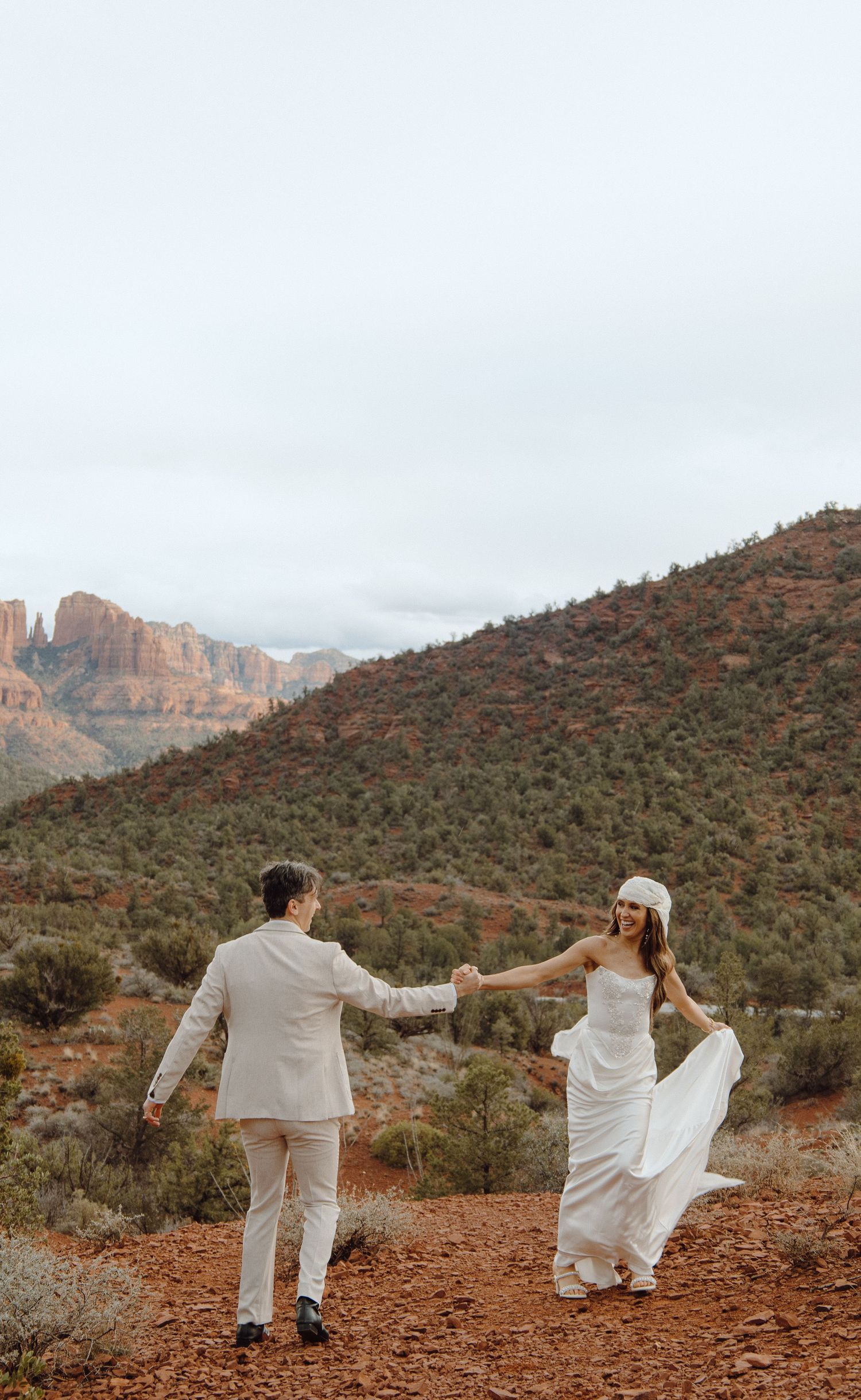 Wedding couple holding hands on a red dirt trail with stunning Sedona red rock formations in the background.