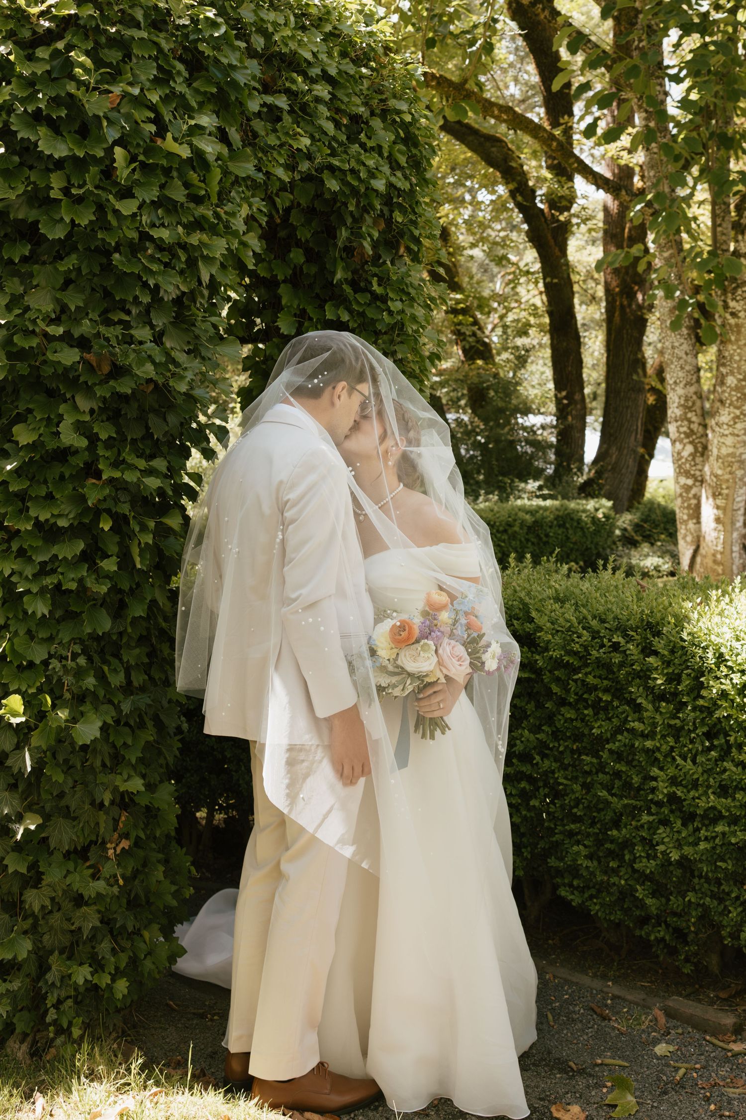 Bride and groom kissing beneath a sheer veil beside an ivy-covered wall, bride holding a colorful bouquet.