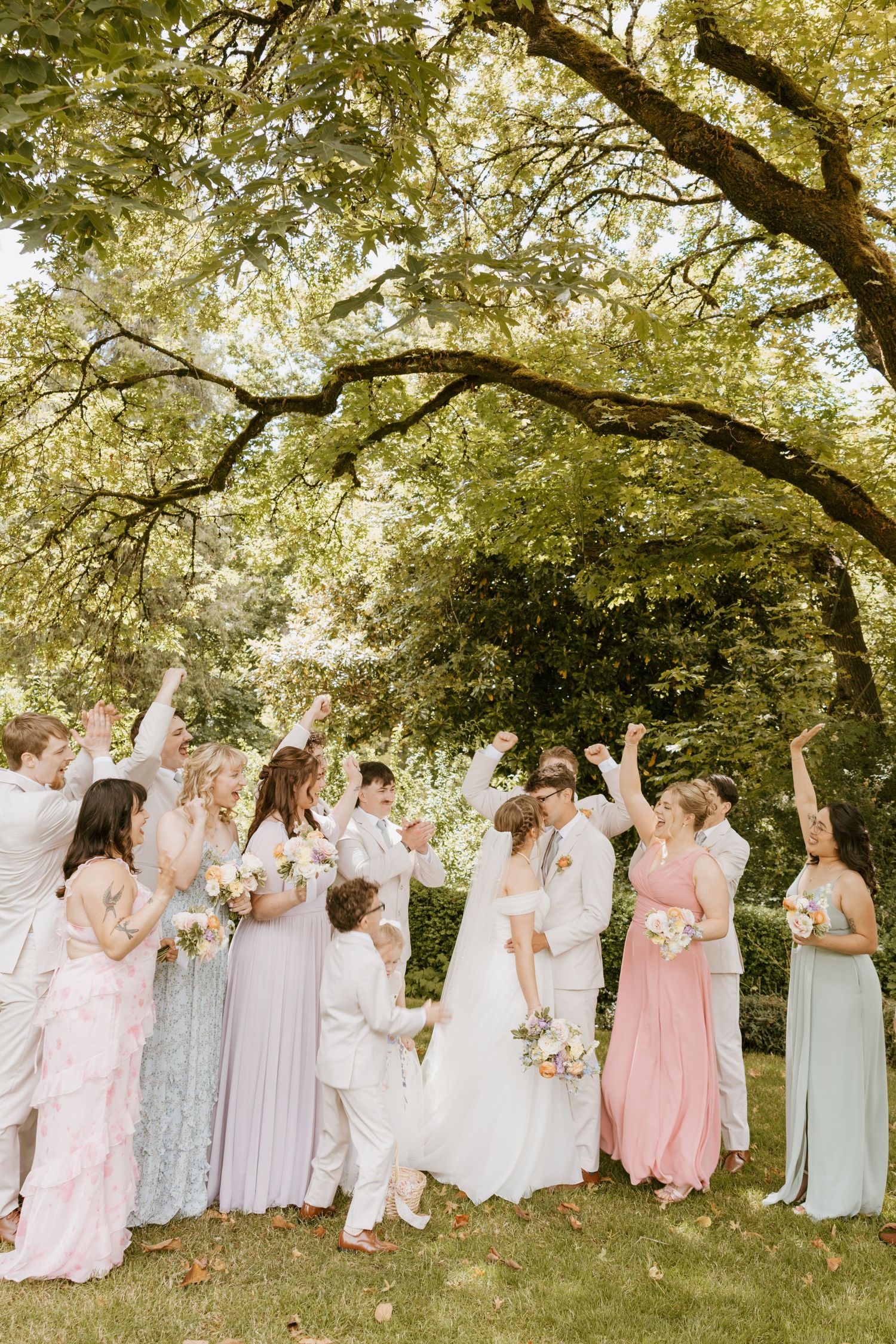 Wedding party celebrating outdoors under large oak tree with bridesmaids in pastel dresses.