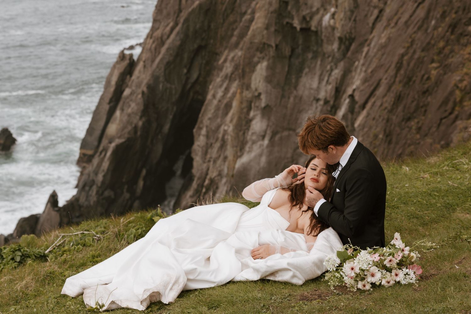 Couple in wedding attire relaxing on cliffside overlooking ocean waves with bridal bouquet nearby.