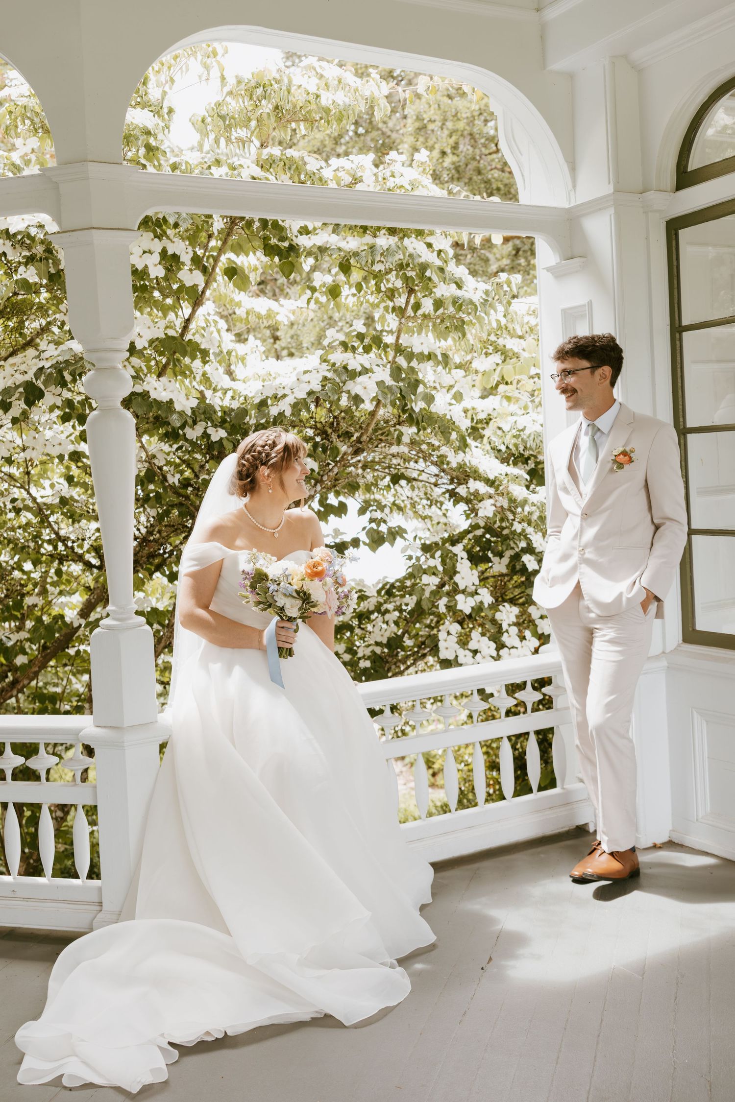 Bride in off-shoulder gown and groom in cream suit share a laugh on sunlit white porch.