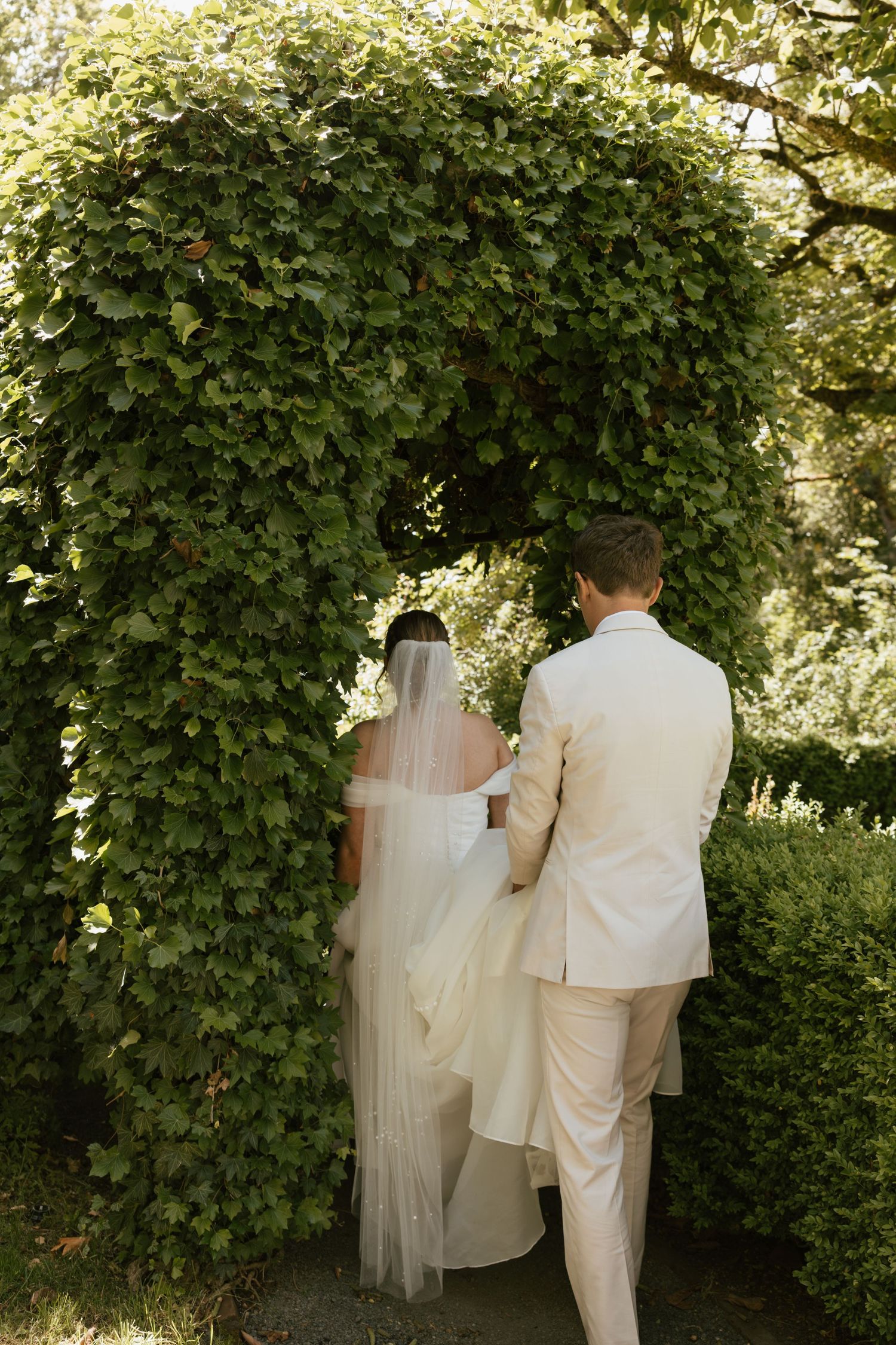 Bride and groom walking hand-in-hand through a lush green ivy-covered archway in a garden setting.