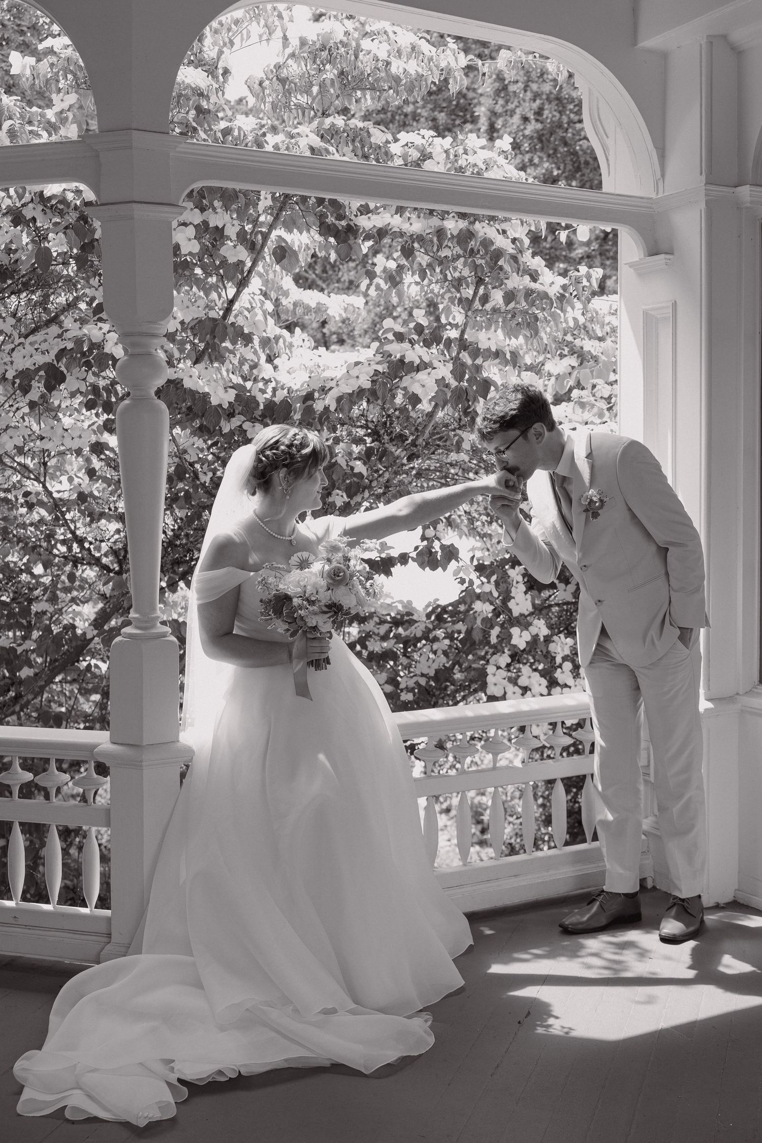 Black and white photo of groom kissing bride's hand on elegant white porch with columns.