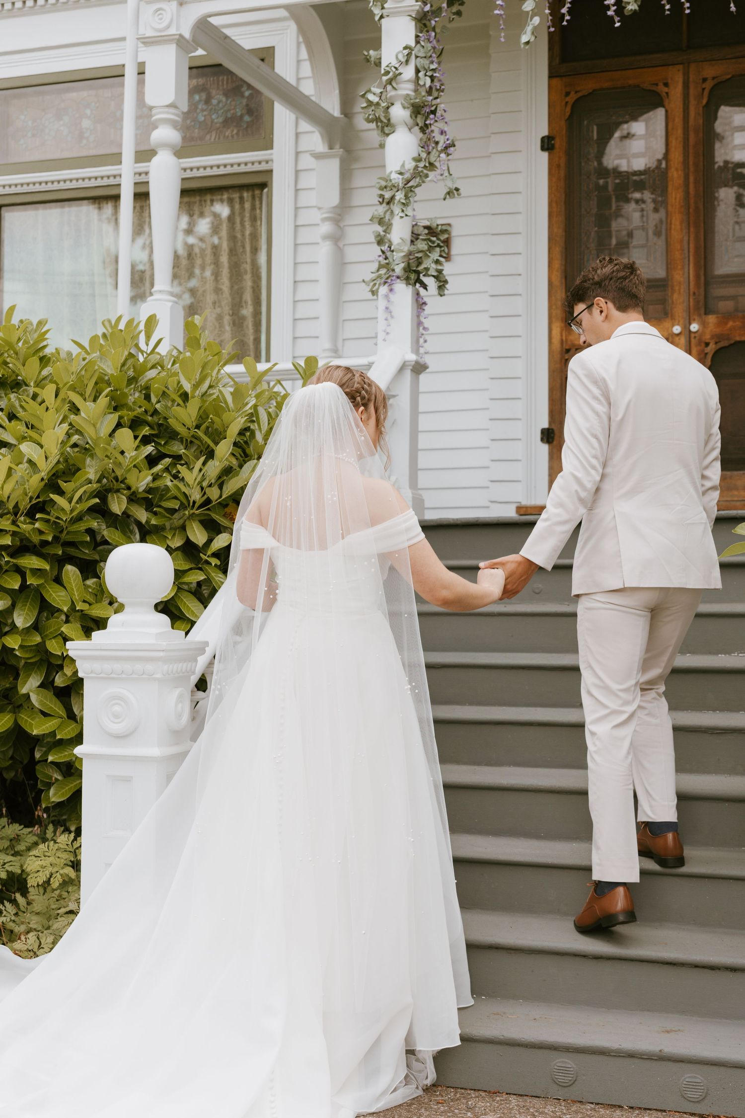 Bride and groom holding hands walking up steps of white Victorian house on wedding day.