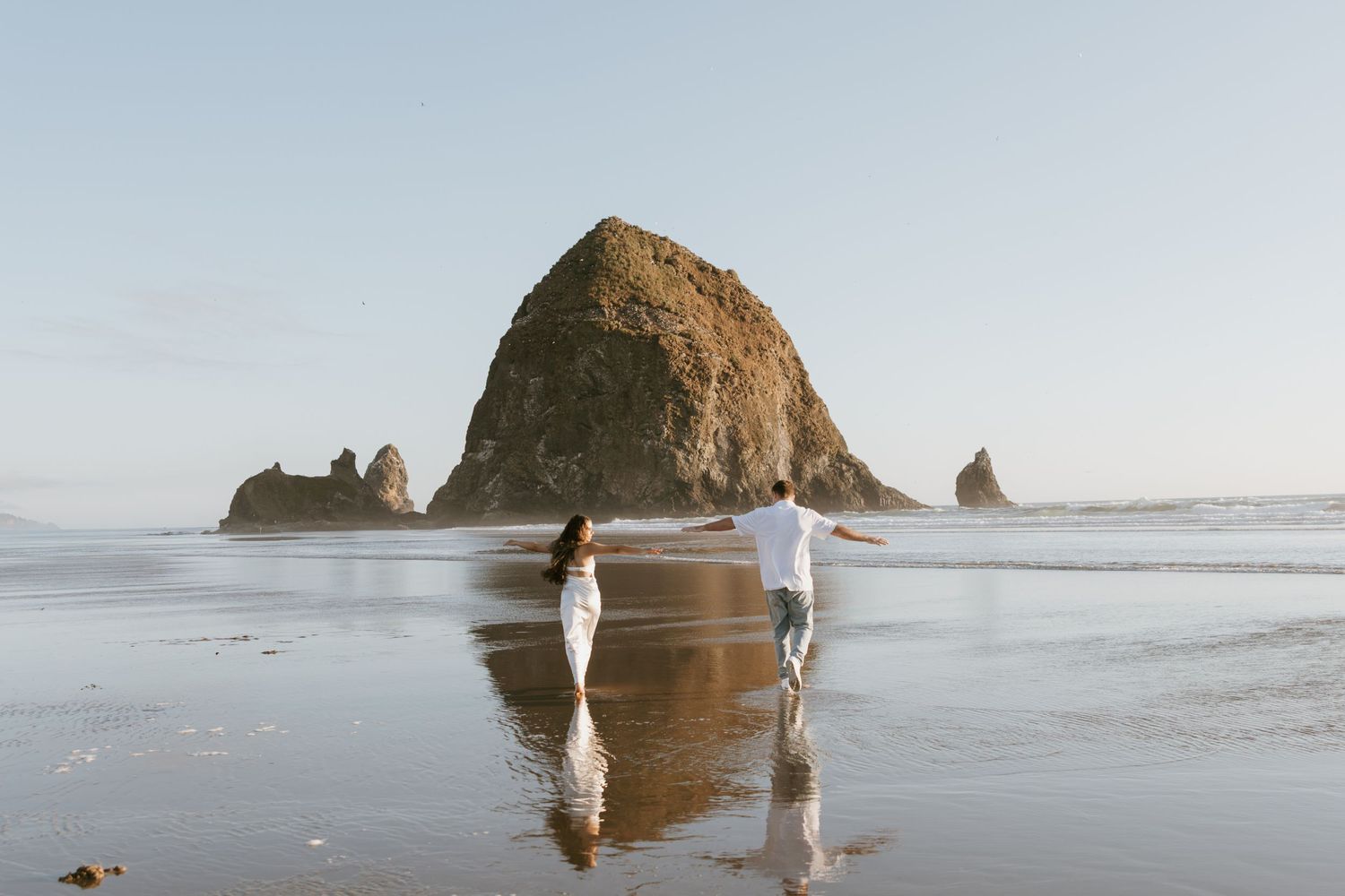 Wedding couple walking hand in hand on reflective beach sand near iconic Haystack Rock in Oregon.
