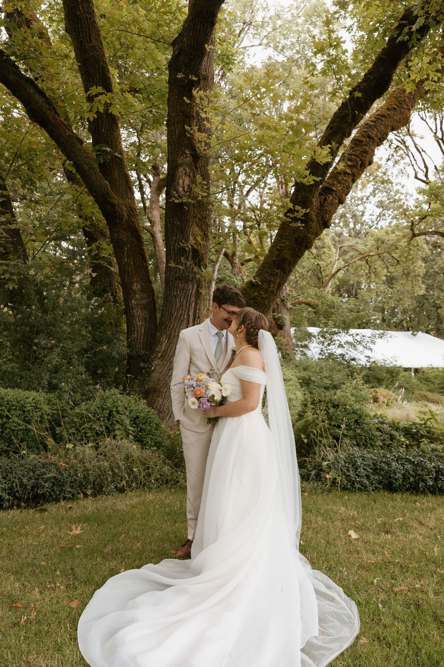 Bride in flowing white gown with veil holds bouquet while kissing groom under large tree.