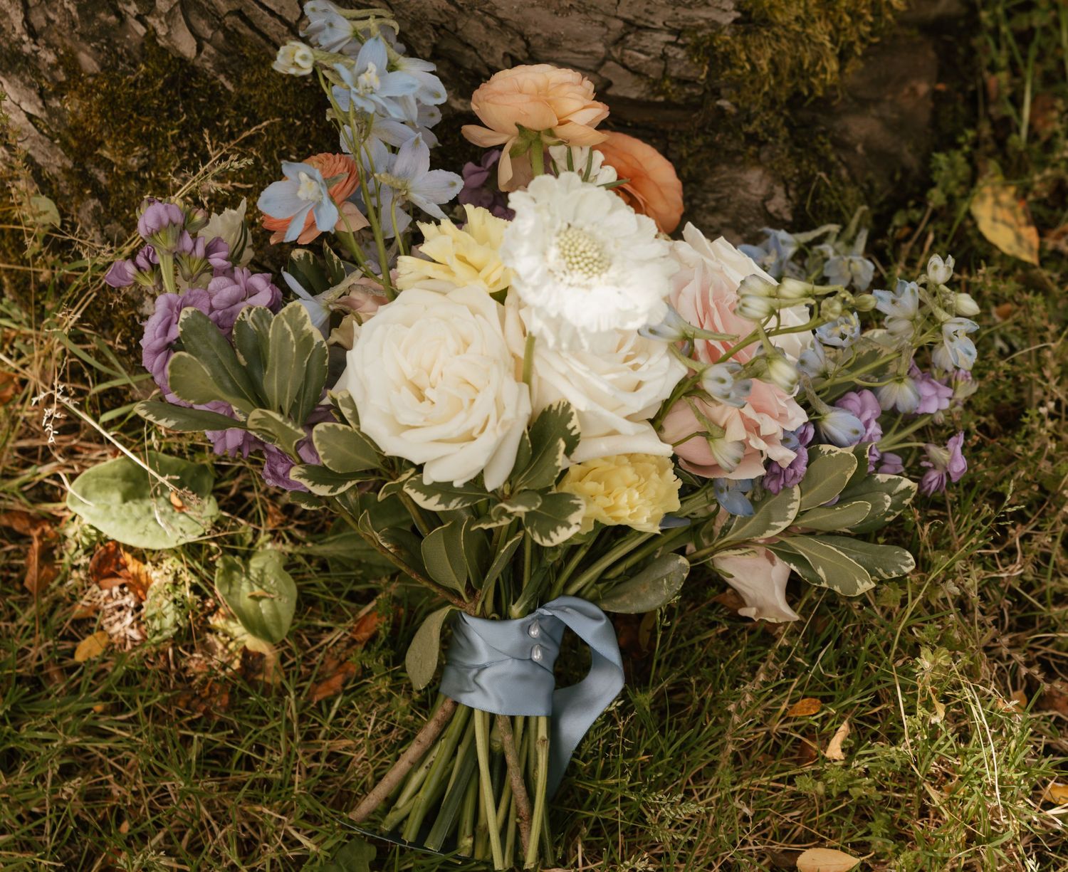 Romantic bridal bouquet featuring white roses, pastel blooms, and blue ribbon on grass.