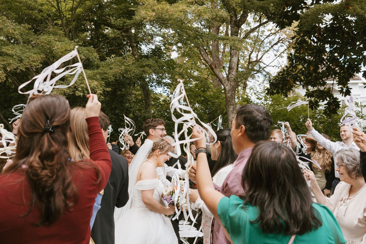 Wedding guests waving ribbon wands as bride and groom walk through a celebratory send-off under tall trees.