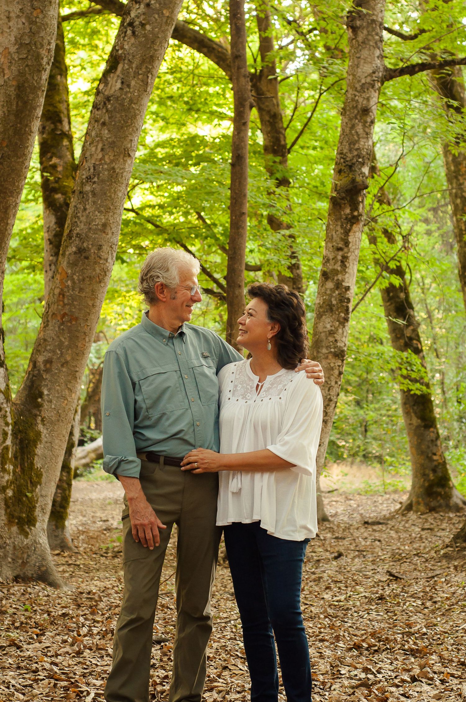 Older couple looking lovingly at each other while standing together among the trees during an outdoor family portrait session.