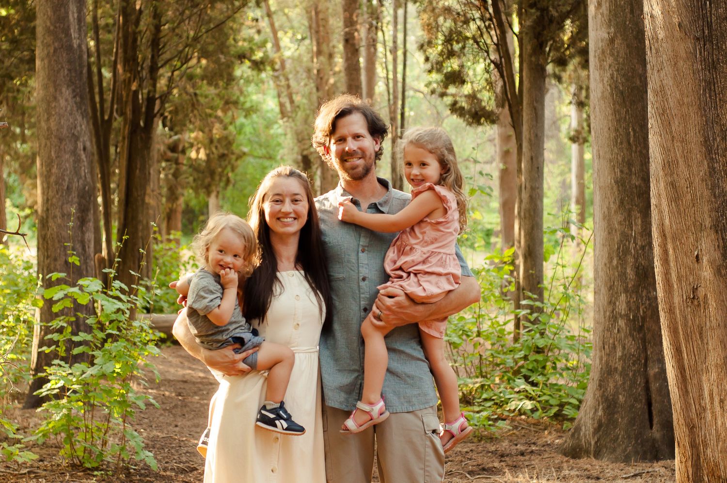 Mother and Father with two small children smiling together among tall trees during a family portrait session.