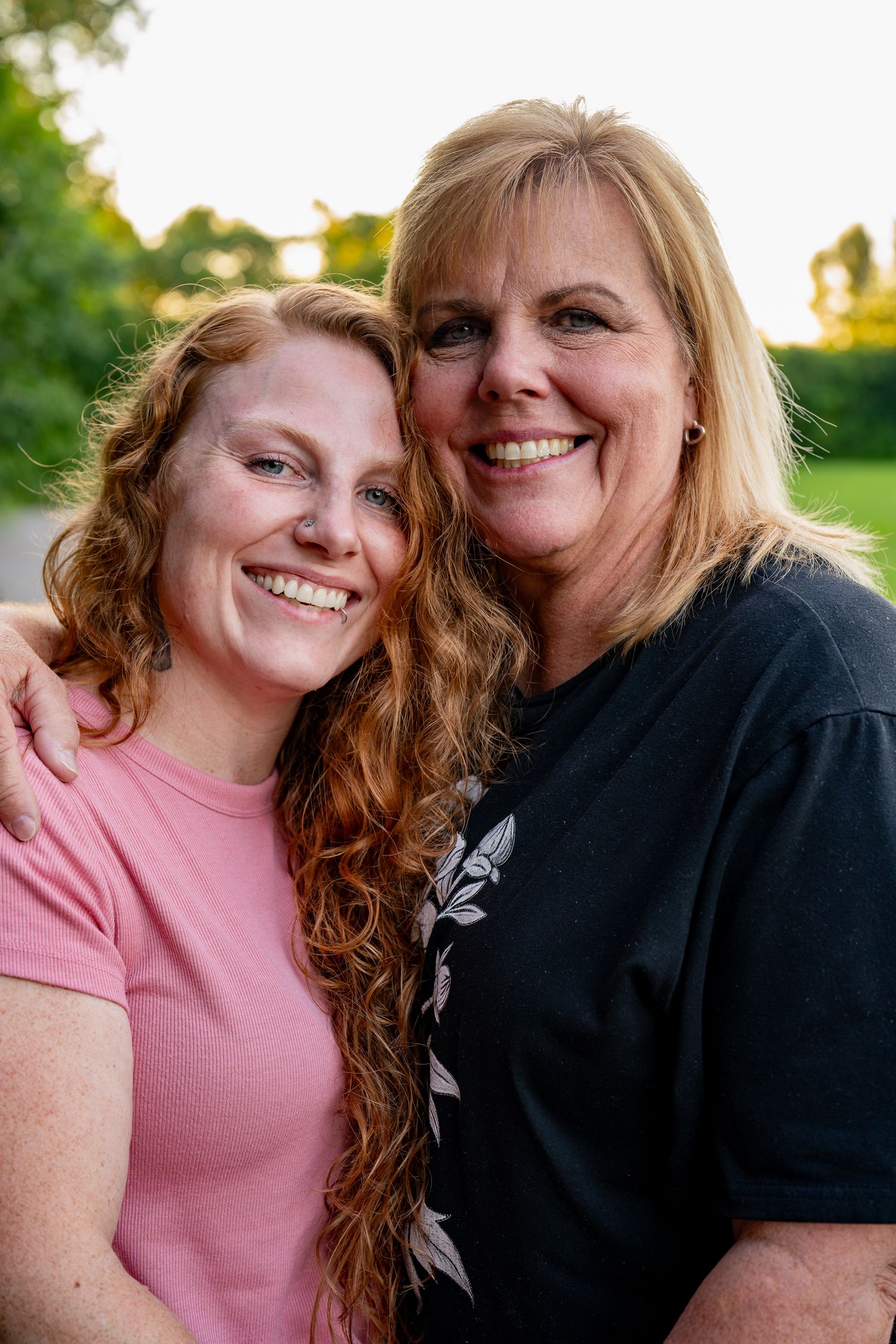 Close-up portrait of a mother and daughter smiling together outdoors at sunset.