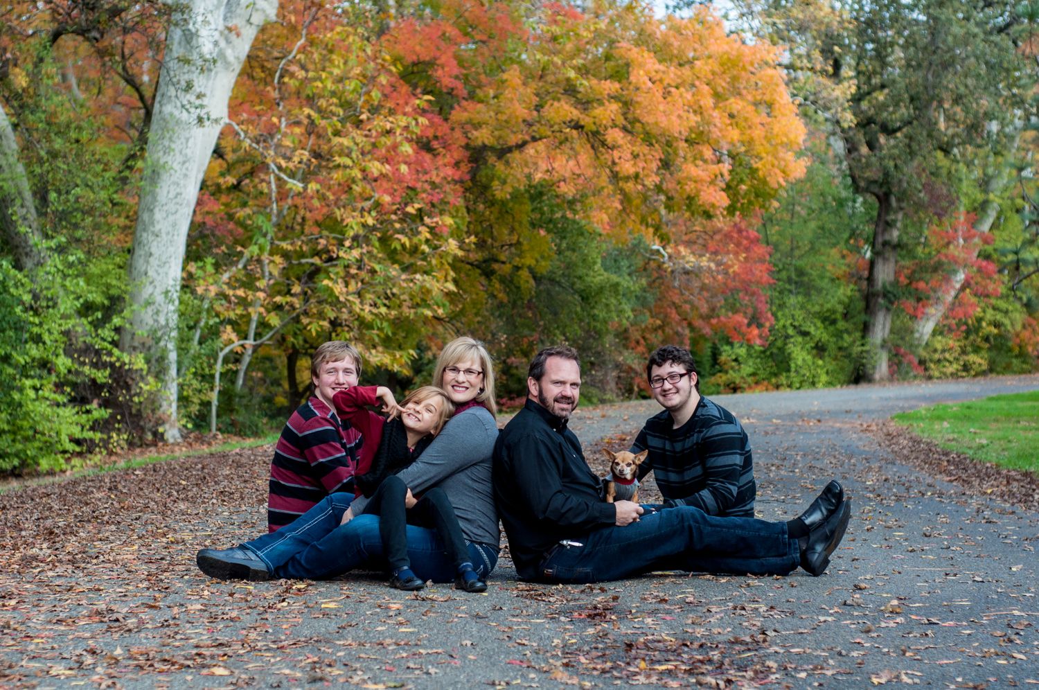 Family sitting together on a tree-lined road surrounded by colorful fall foliage.