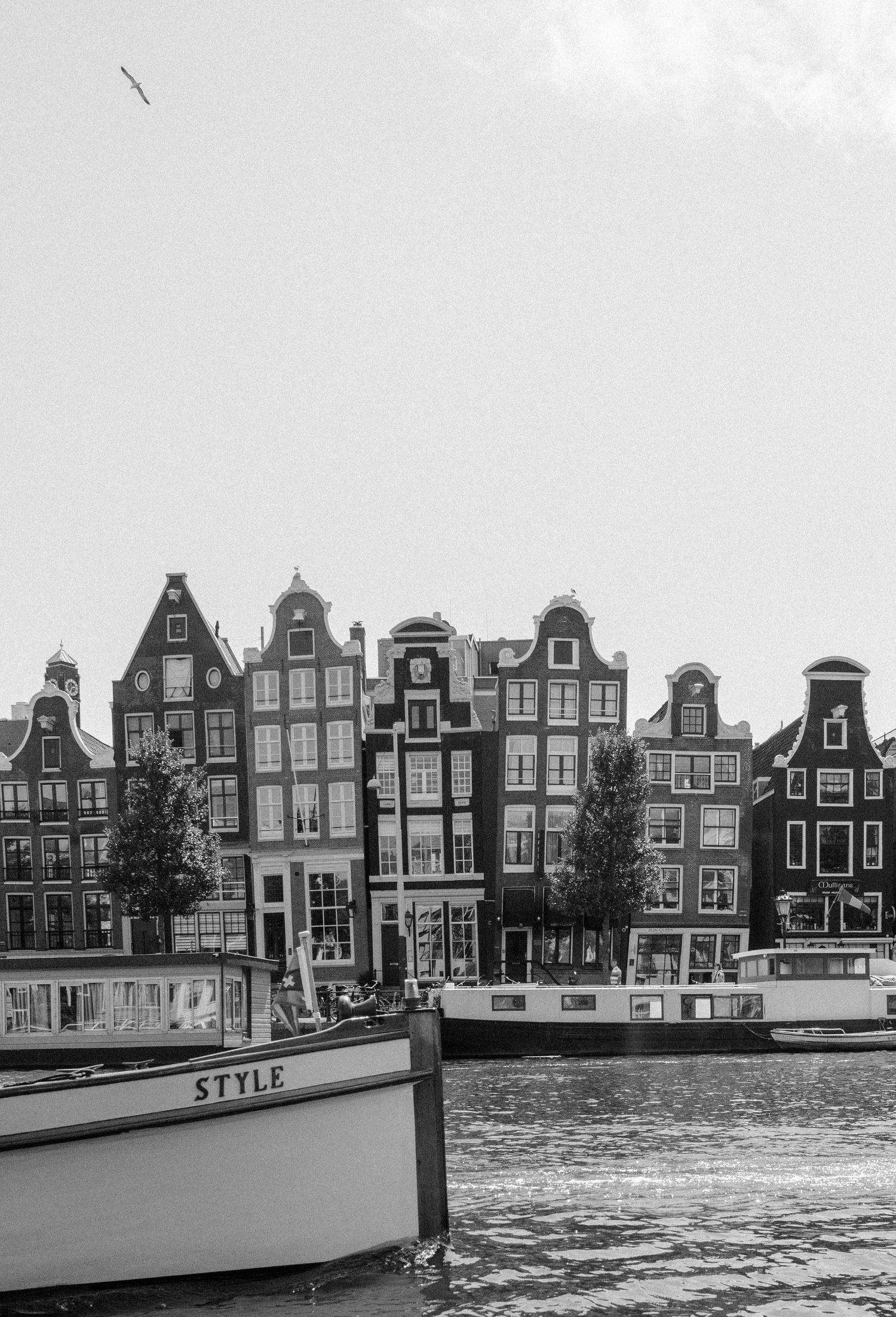 Black and white image of a boat sailing in a canal with buildings in the background in Amsterdam