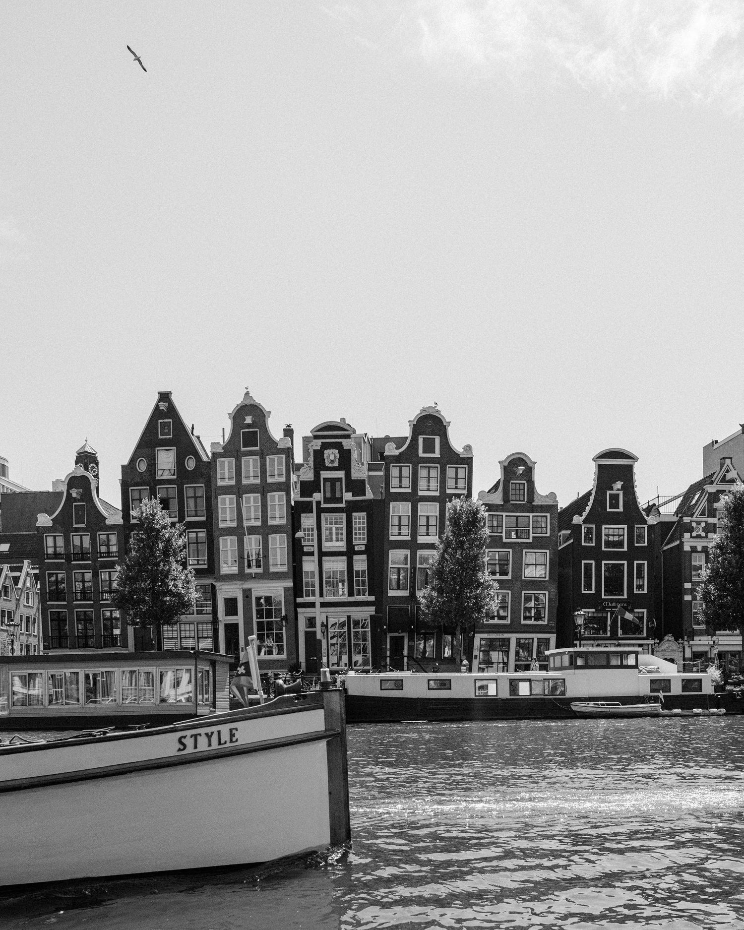 Black and white image of small boat sailing in the water of a canal in front of buildings in Amsterdam.
