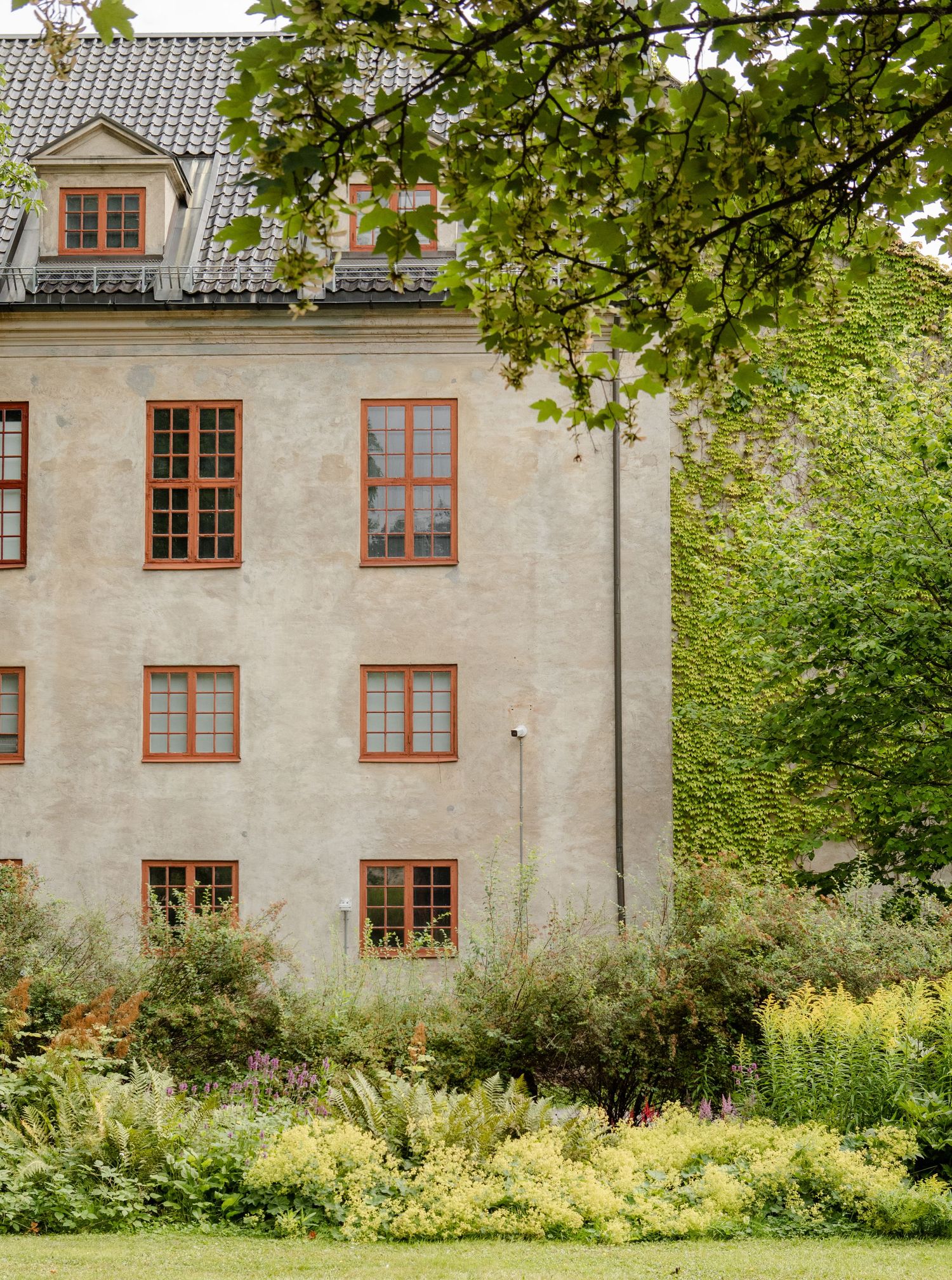 A building with red bordered windows and grey stone wall among greenery, trees and grass in Norway
