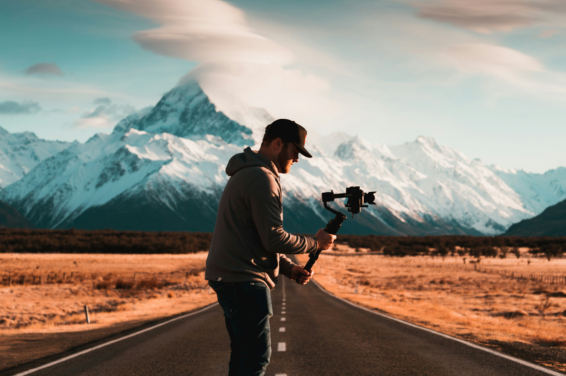 Man standing in the middle of a road with a mountain beyond him.  Man is filming nature on the east side of the road.