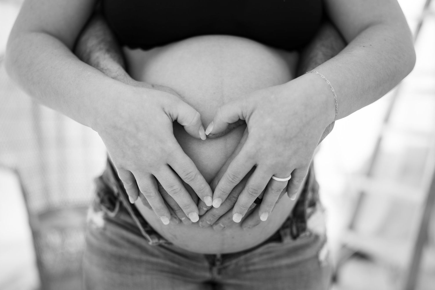 Black and white close-up of hands making heart shape on expectant mother's bare belly.
