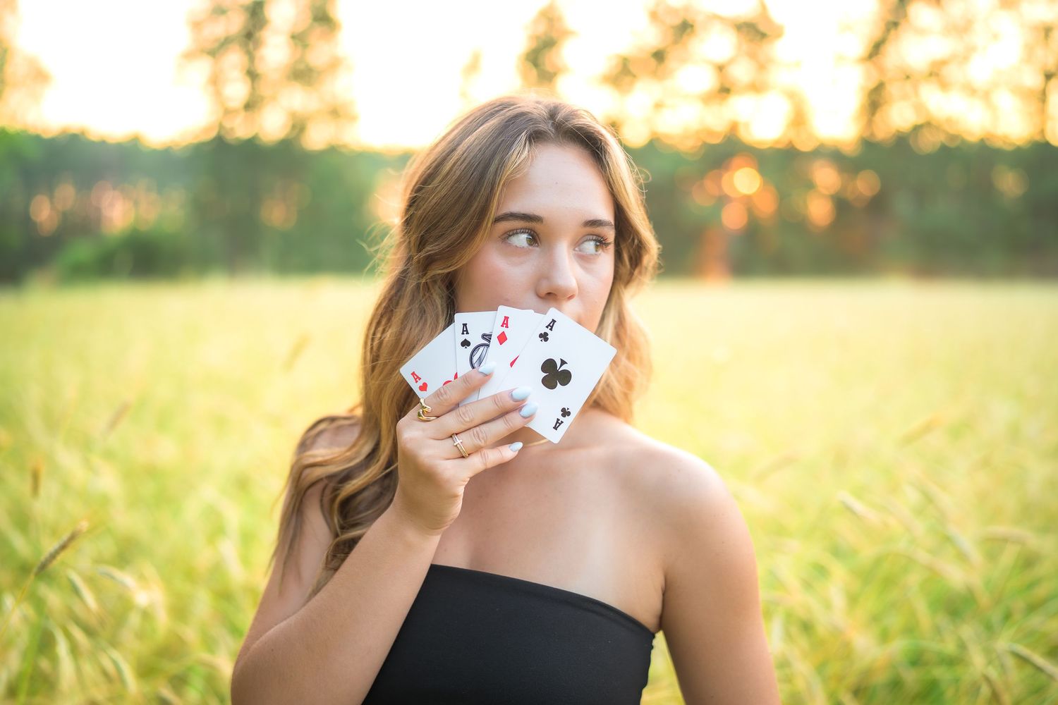 Someone in a black top holds playing cards in front of their face while sitting in a golden sunset field.