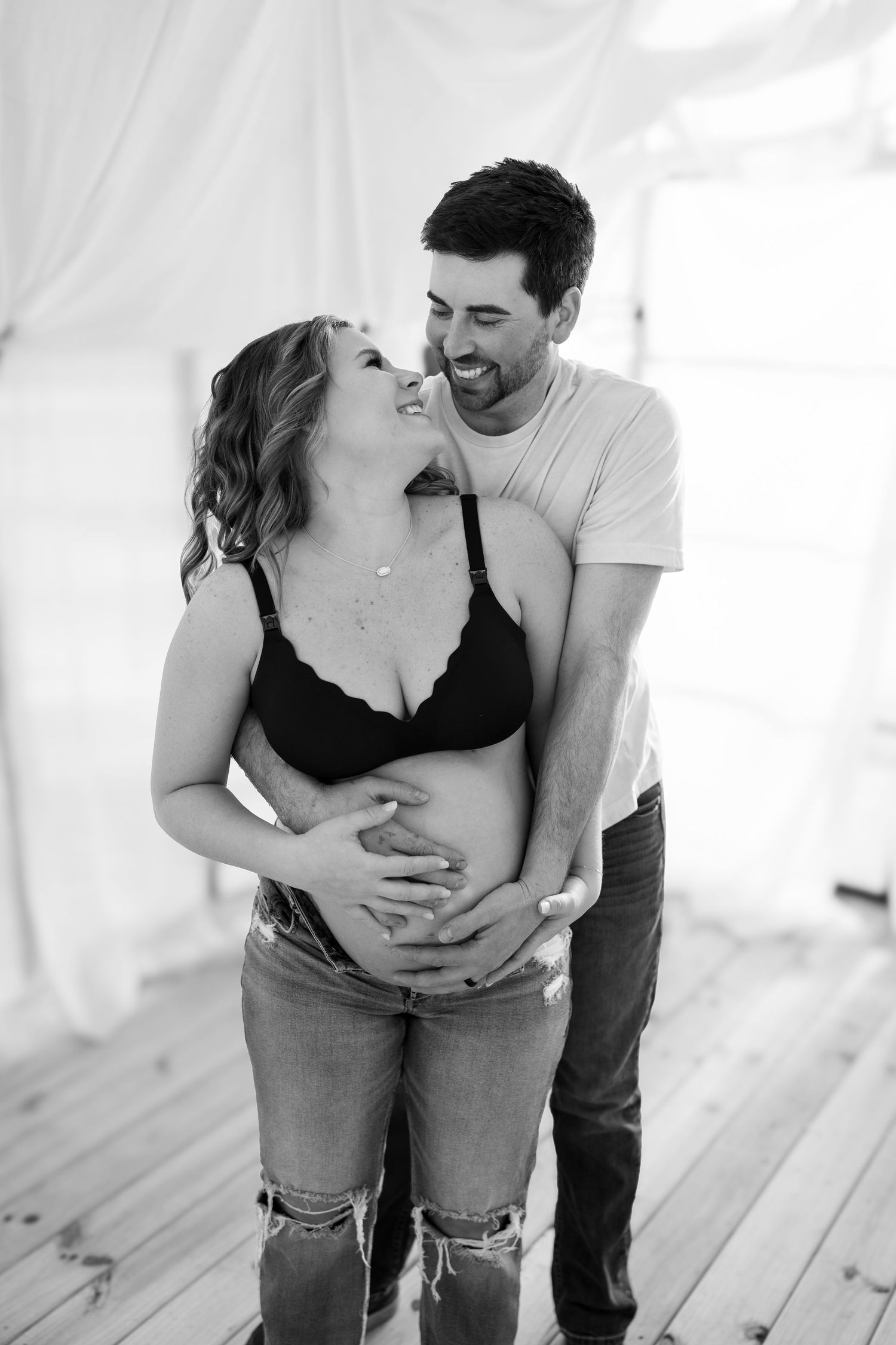 Black and white maternity photo showing couple laughing together while holding pregnant belly.