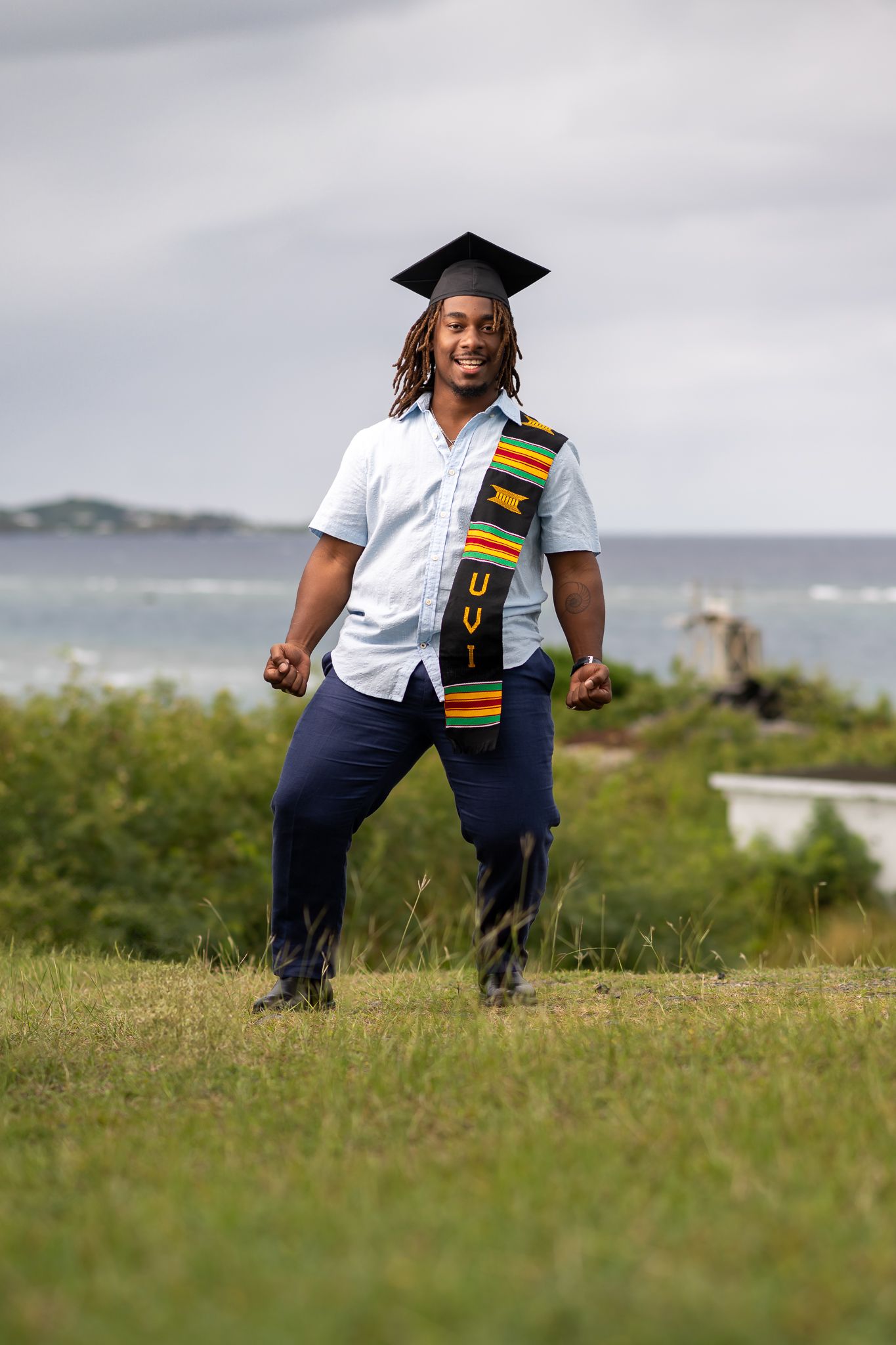 Graduate in white shirt and navy pants poses with a cultural stole against coastal landscape.